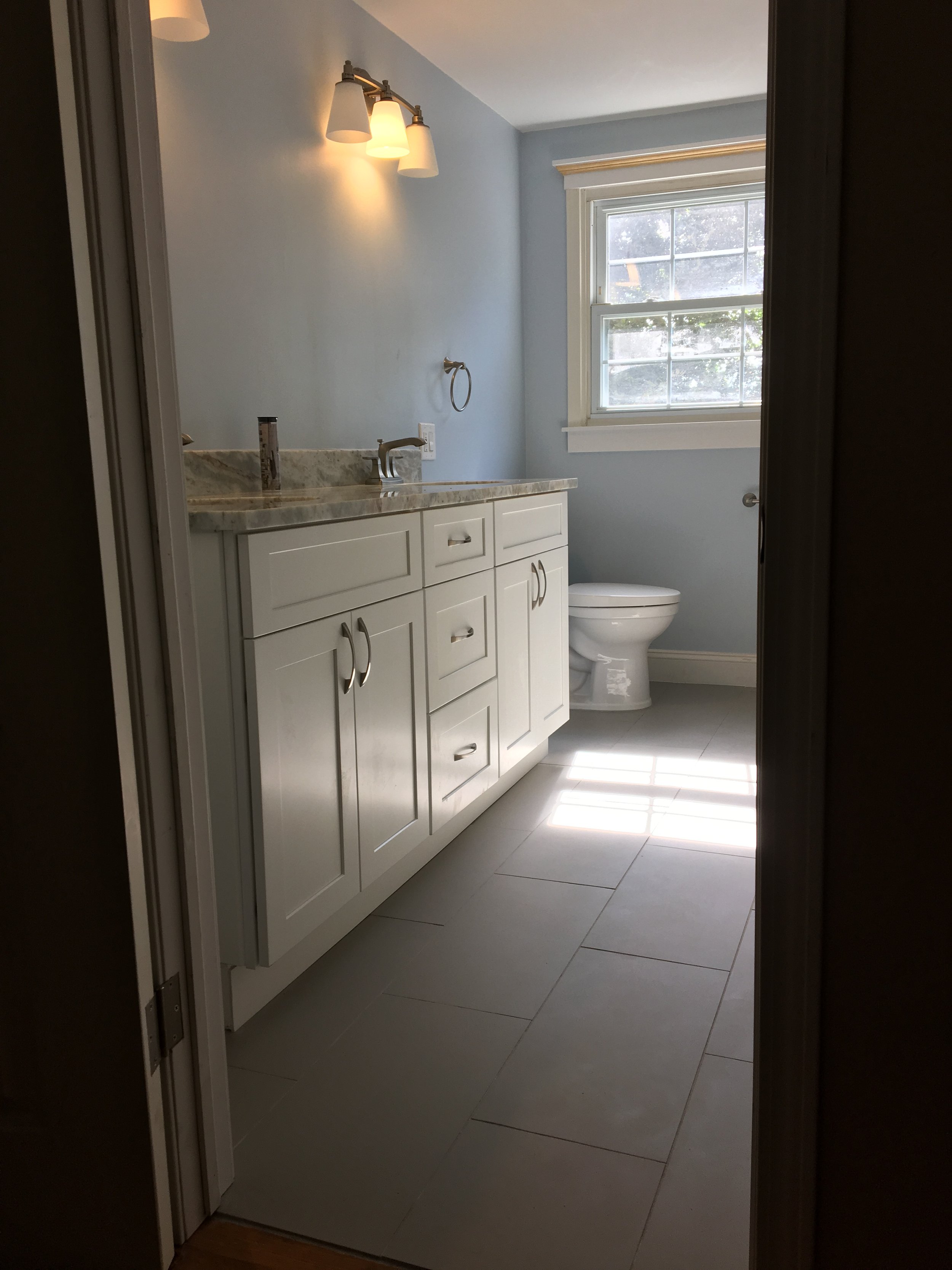 A bathroom with a white vanity, a marble countertop, a sink with a silver faucet, a wall-mounted mirror, a light fixture with three lights, a towel ring, a toilet in front of a window with white trim, and beige tiled flooring.
