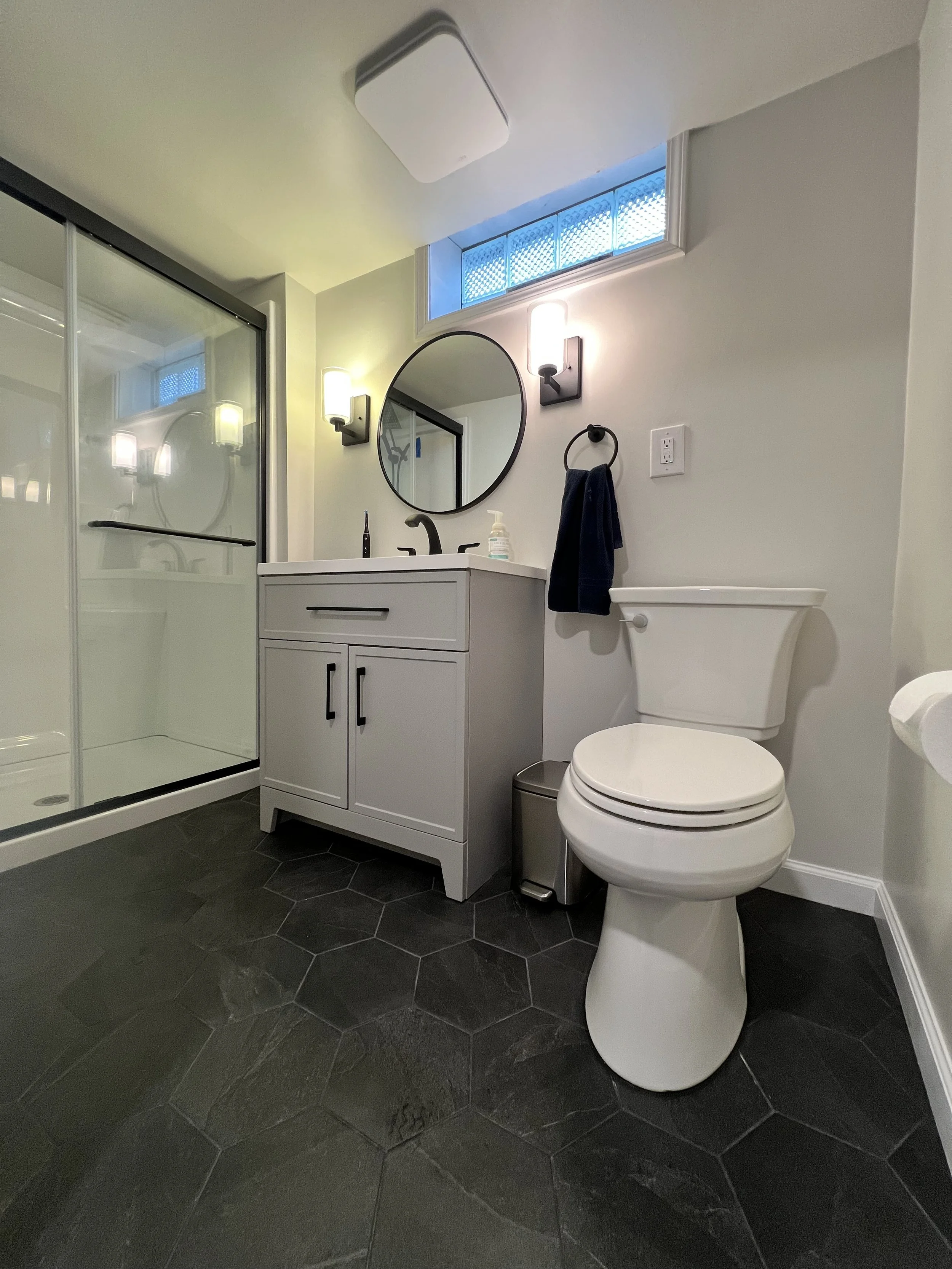 Modern bathroom with a walk-in shower, vanity with a round mirror, and a toilet. The floor is black hexagonal tiles, and there's a small high window with blue glass blocks.