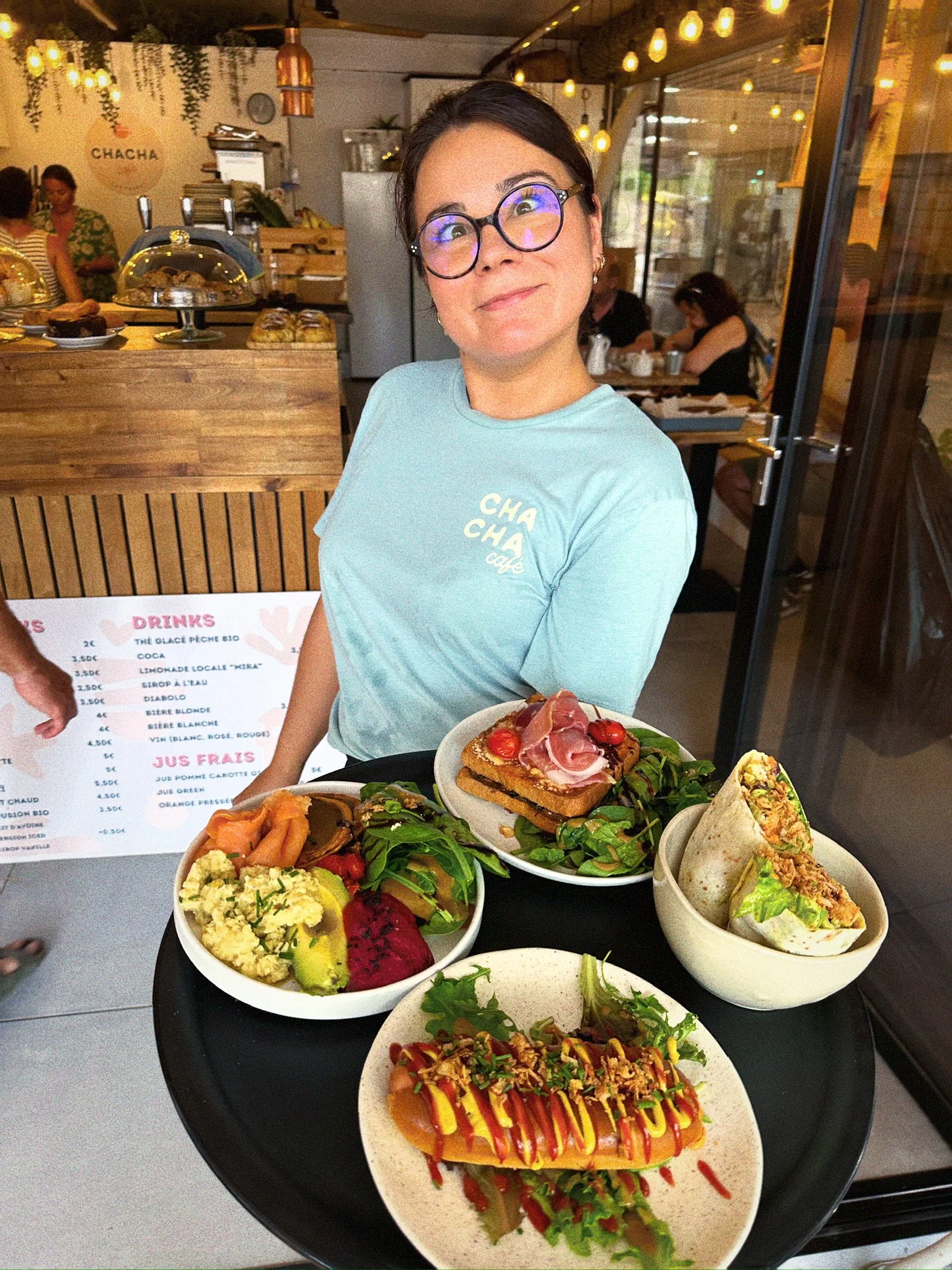Femme souriante portant un t-shirt bleu clair avec le logo 'CHA CHA Cafe', servant des plats végétariens et végétaliens dans un café lumineux avec des décorations chaleureuses.
