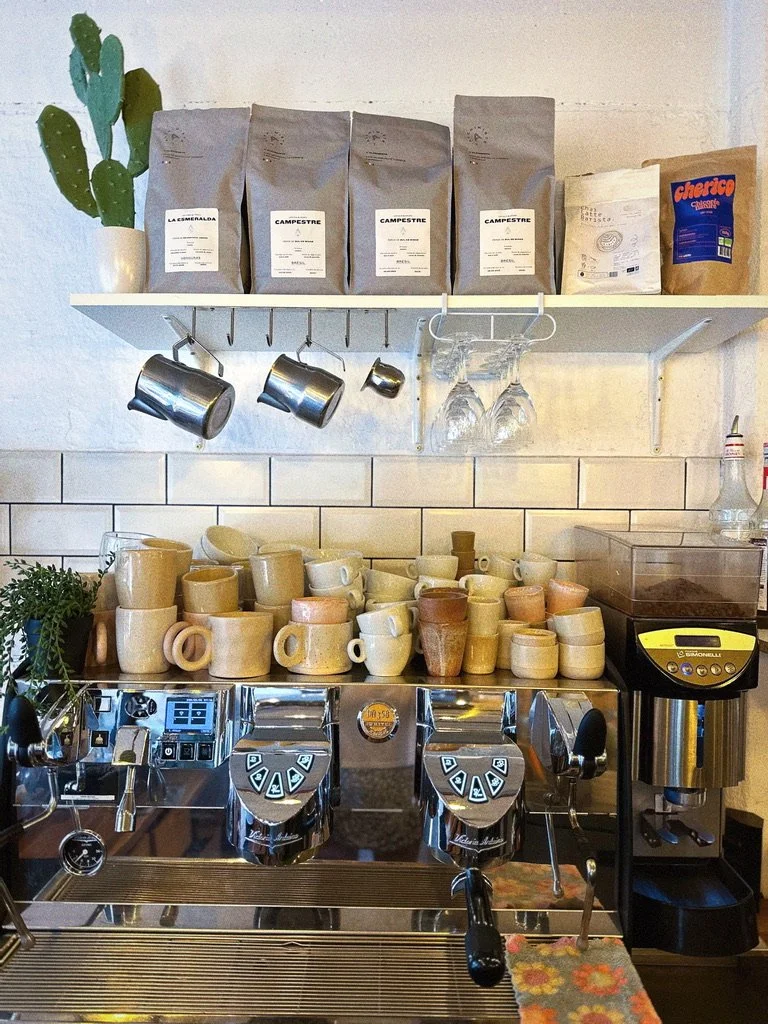 Étal de café avec plusieurs mugs en céramique, une machine à espresso, des sacs de café, et des verres suspendus.