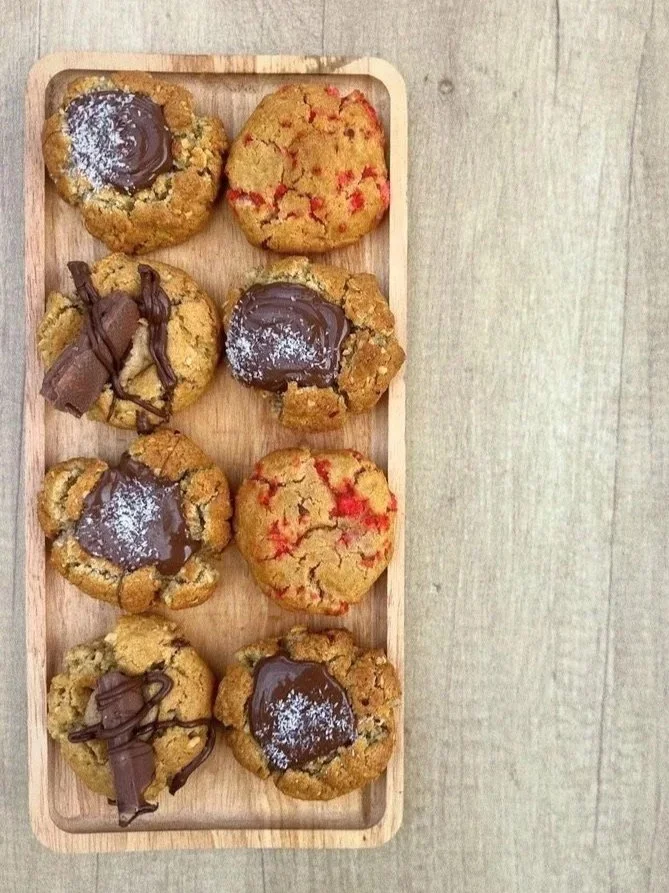 Plateau en bois avec six cookies aux pépites de chocolat, certains décorés de chocolat fondu ou en spirale, et d'autres saupoudrés de sucre glace, étalé sur une surface en bois ou en pierre.