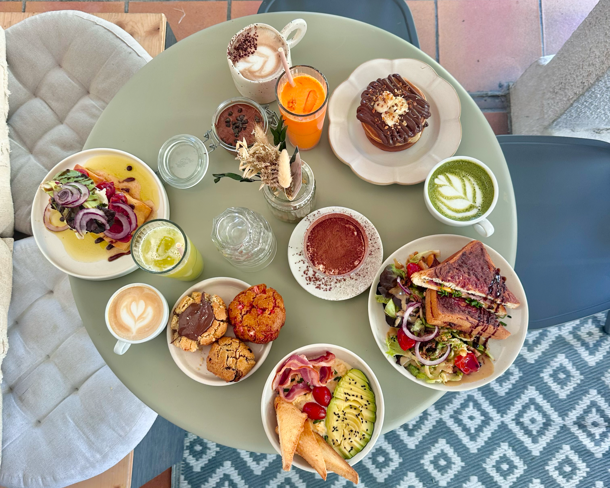 Petit-déjeuner ou brunch avec différentes assiettes de nourriture et boissons, comprenant une salade, des sandwichs, des pâtisseries, des smoothies, et des cafés, sur une table ronde verte.