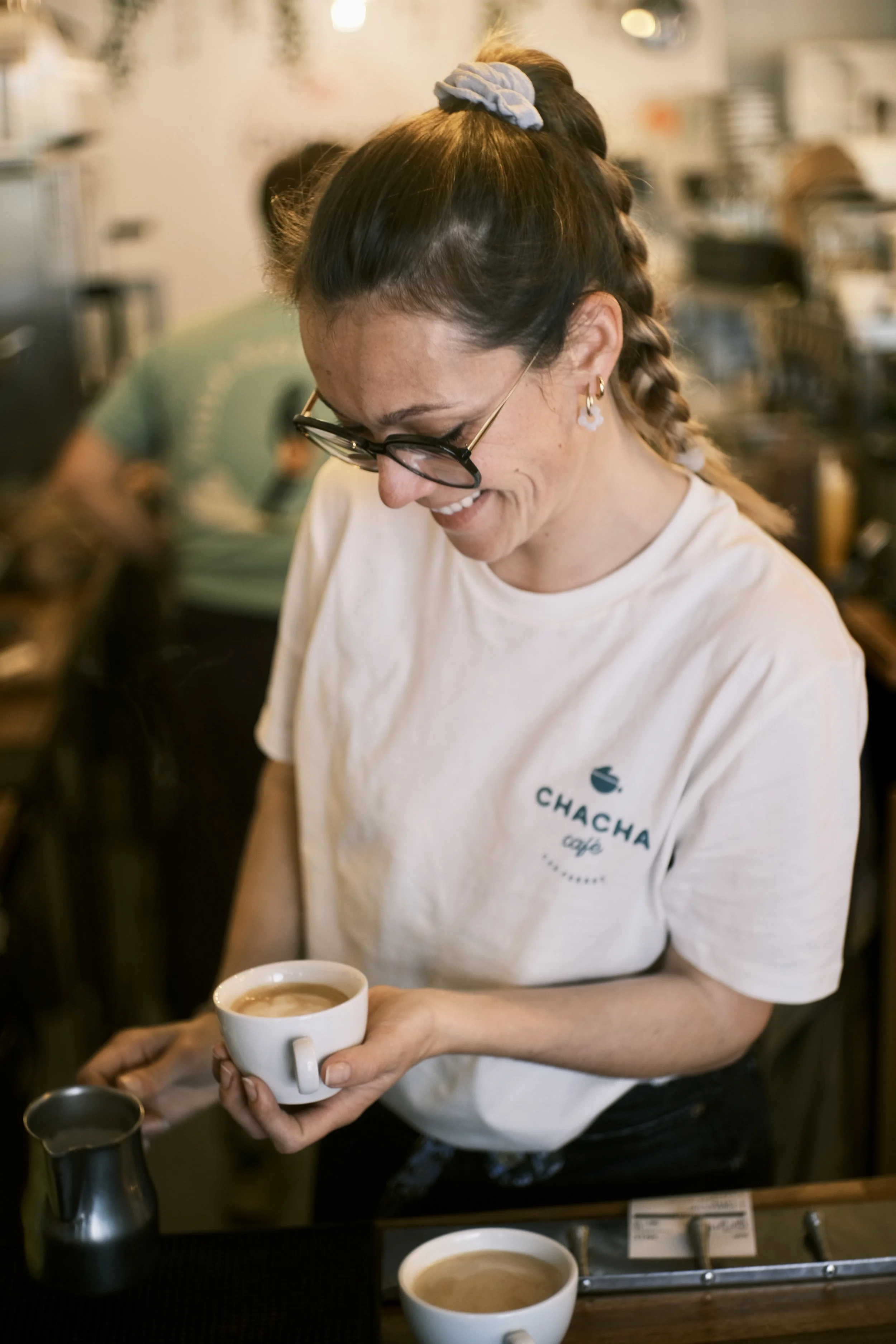 Une barista souriante tient une tasse de café dans un café nommé Chacha, avec un décor chaleureux en arrière-plan.