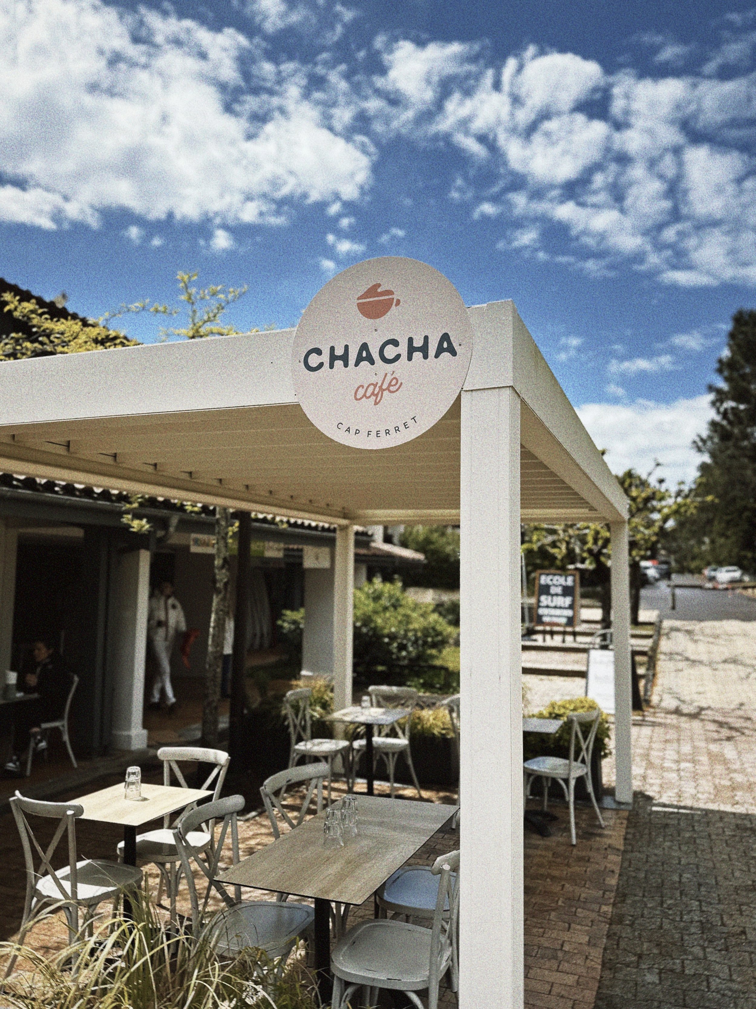 Extérieur d'un café nommé Chacha avec un panneau rond montrant un logo de tasse de café, des tables et des chaises en bois, un ciel bleu avec quelques nuages, dans un cadre décontracté et en plein air.