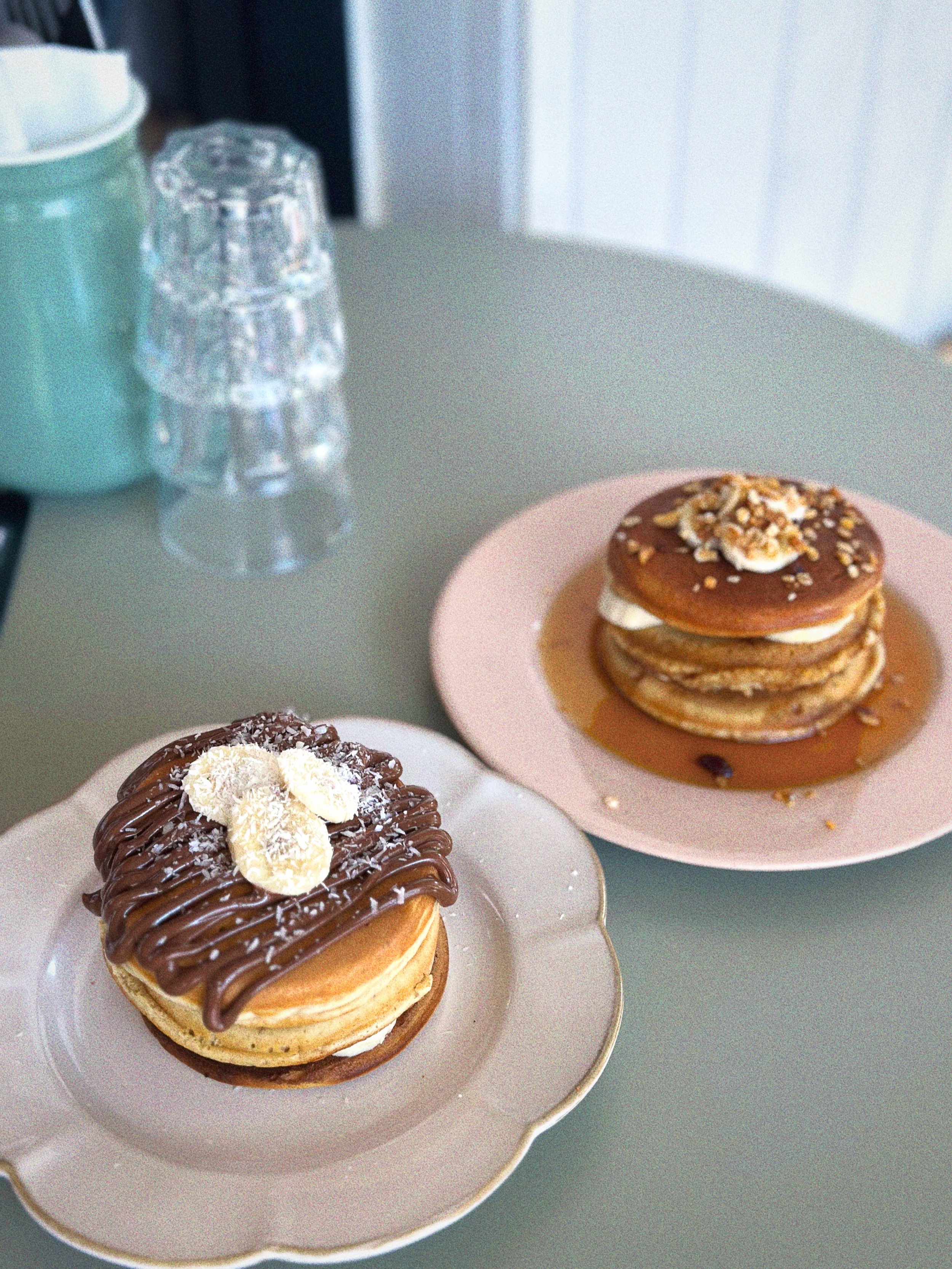 Deux desserts de pancakes garnis, un avec du chocolat et des copeaux de noix de coco, l'autre avec du caramel, des noix et de la crème, sur deux assiettes différentes.