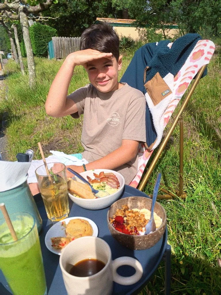 Jeune garçon assis à une table en plein air avec des plats et des boissons, saluant du revers de la main.