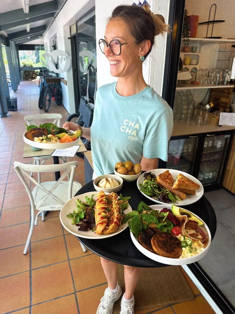 Une femme souriante tenant un plateau avec plusieurs plats de nourriture, dans un café ou restaurant, avec une ambiance intérieure chaleureuse et lumineuse.