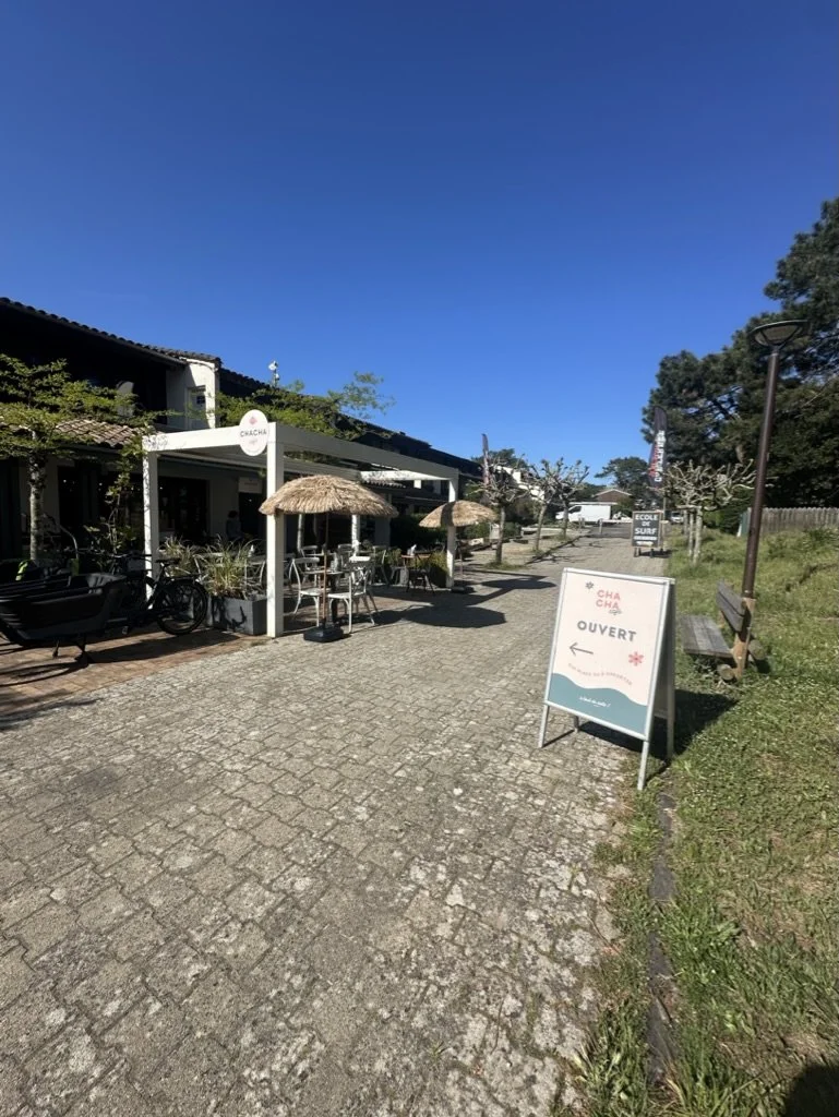 Une terrasse de café avec des tables et des parasols en paille, un bâtiment en arrière-plan, un chemin pavé, et un panneau indiquant que l'endroit est ouvert, dans un environnement en plein air avec des arbres et un ciel bleu clair.