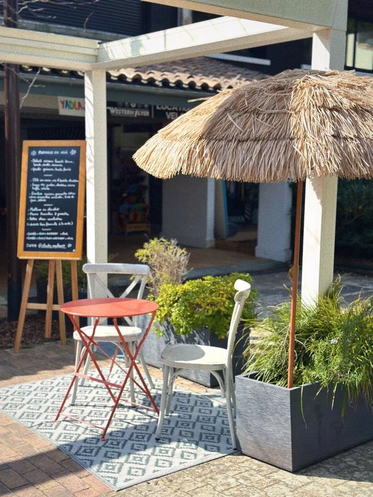 Un coin extérieur d'un café ou restaurant avec une petite table rouge, deux chaises, un parasol en paille, des plantes en pot et un panneau au fond.
