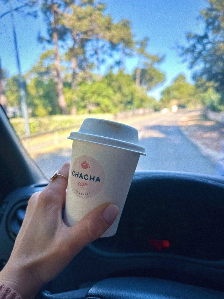 Une personne tient une tasse de café blanche avec un logo 'Chacha' à l'intérieur d'une voiture, vue d'une route en dehors avec des arbres verts et un ciel bleu.