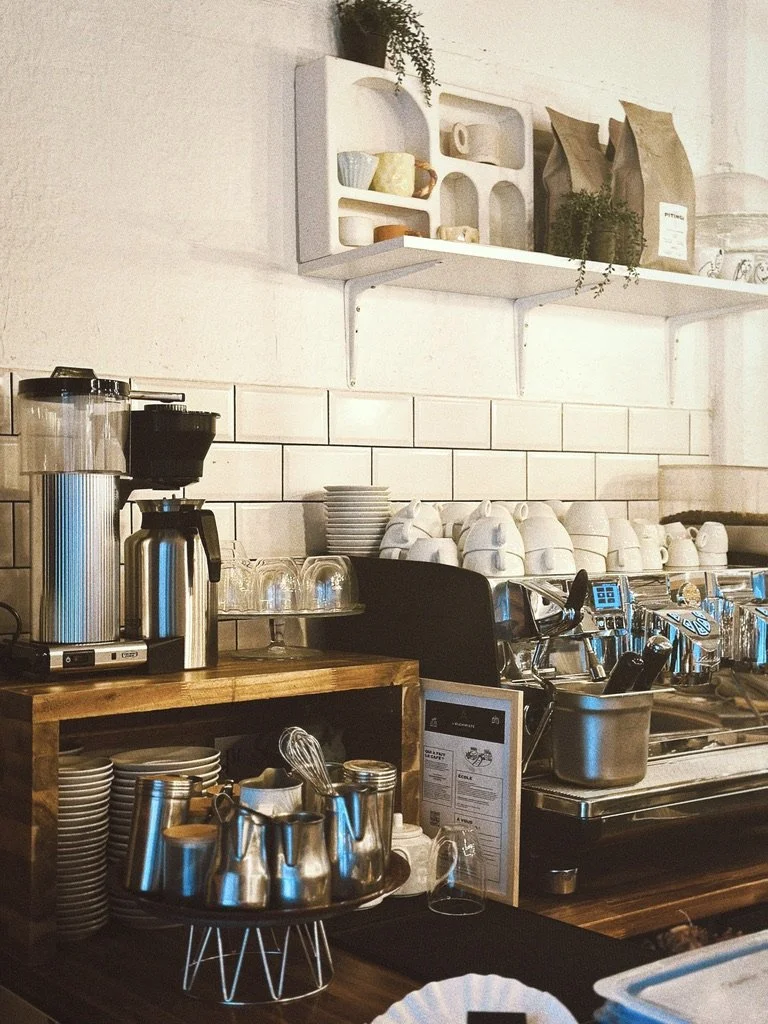 Coins, mugs, and utensils on a wooden countertop in a café kitchen, with a coffee machine and cups in the background.