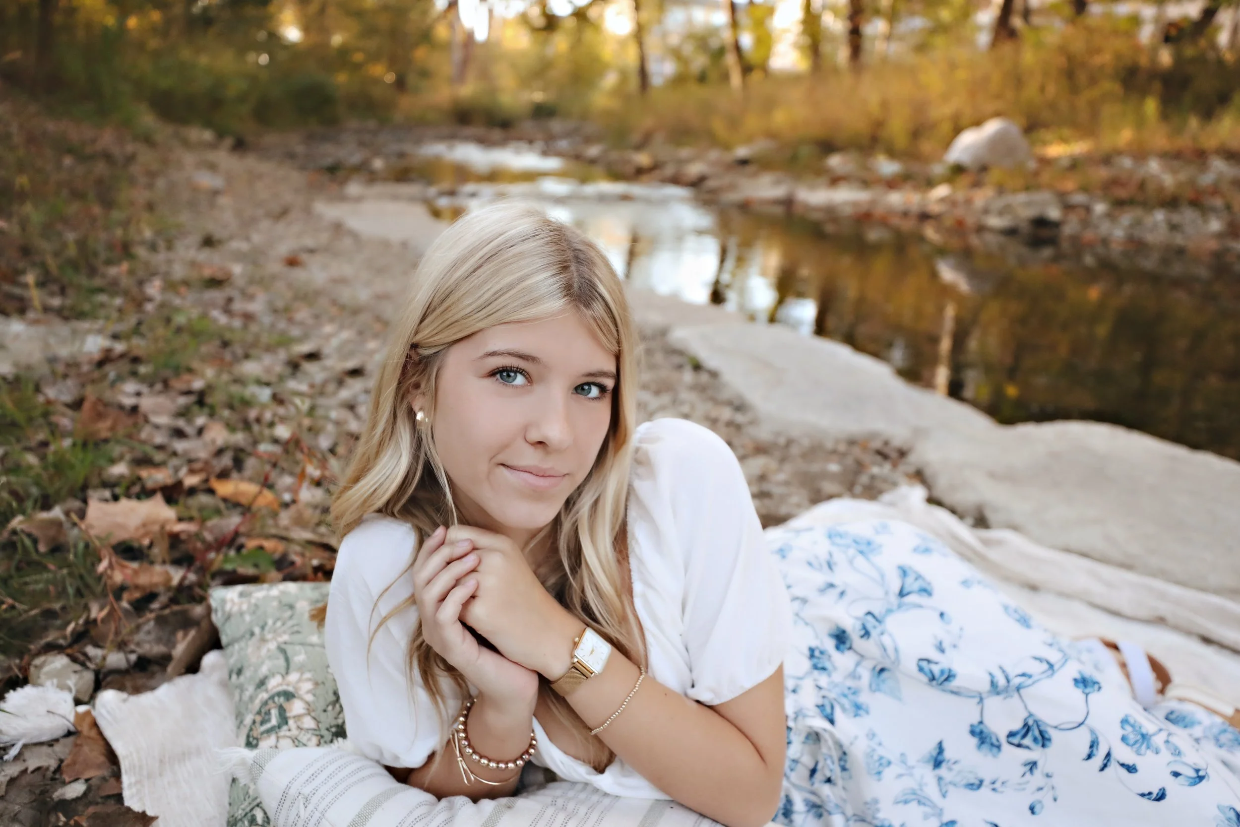 A young woman with blonde hair and blue eyes lying on a blanket near a creek in autumn.