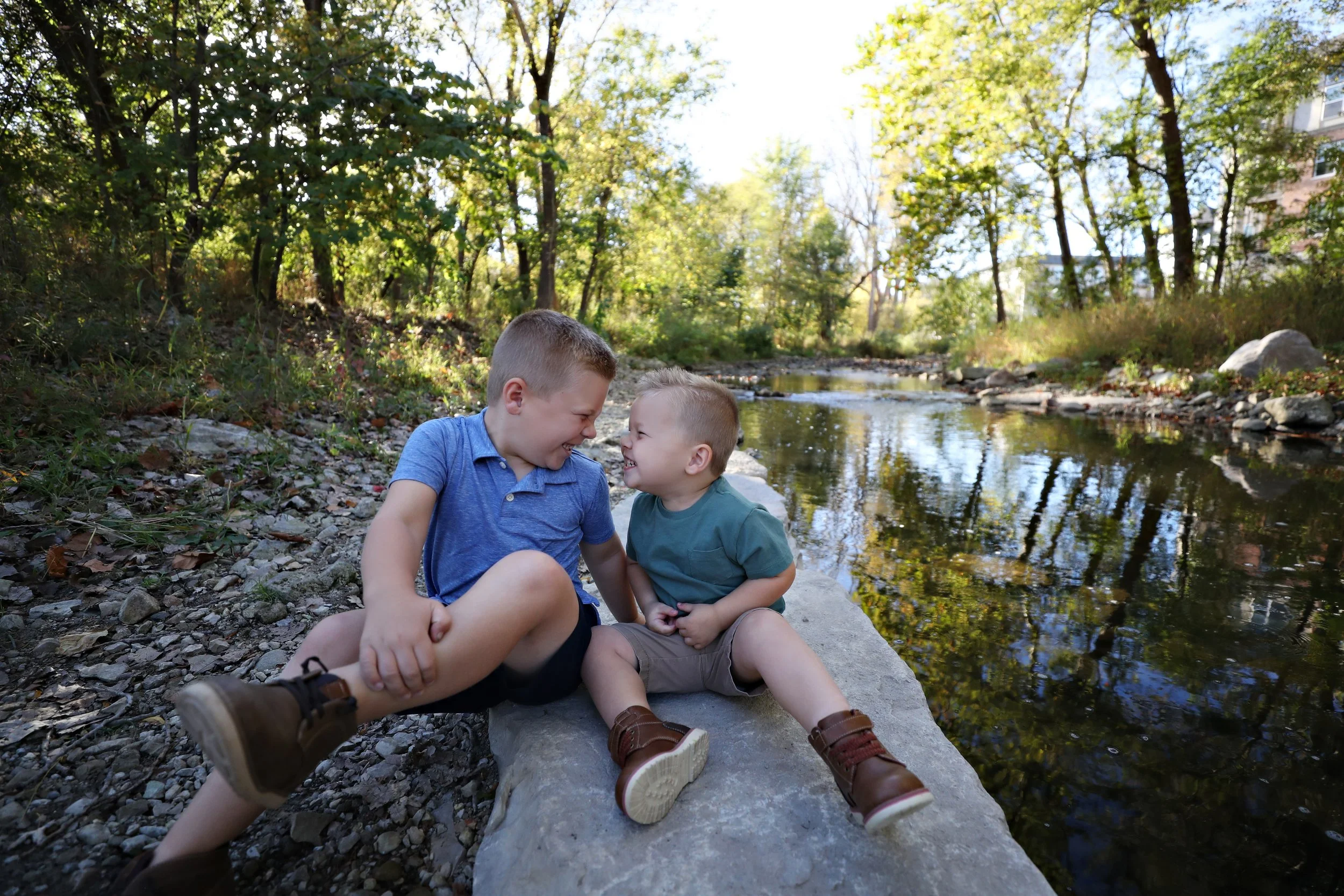 Two young boys smiling and laughing while sitting on a large rock by a creek surrounded by trees.