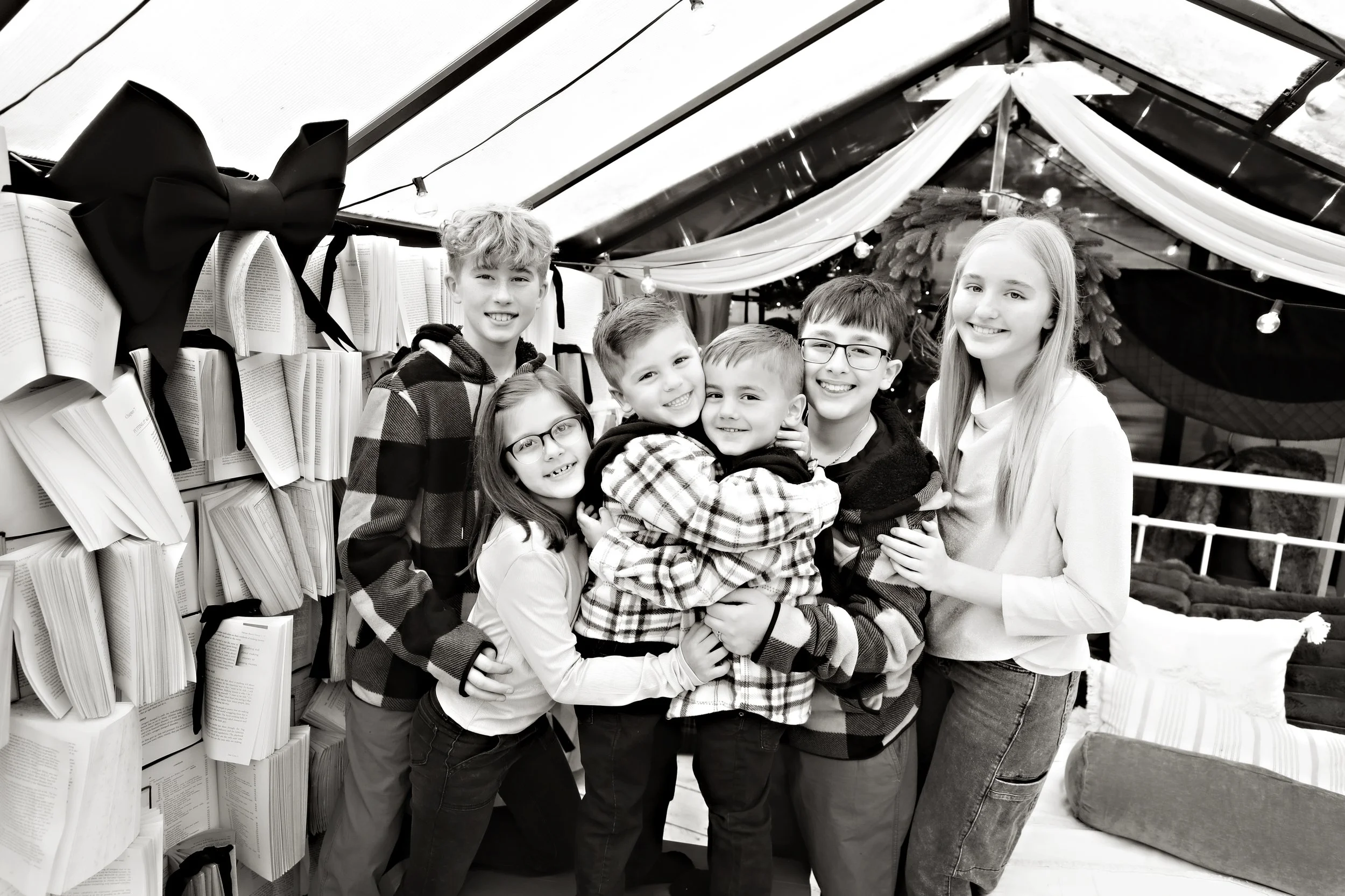 Group of seven children, smiling and hugging, inside a decorated tent with books and string lights.