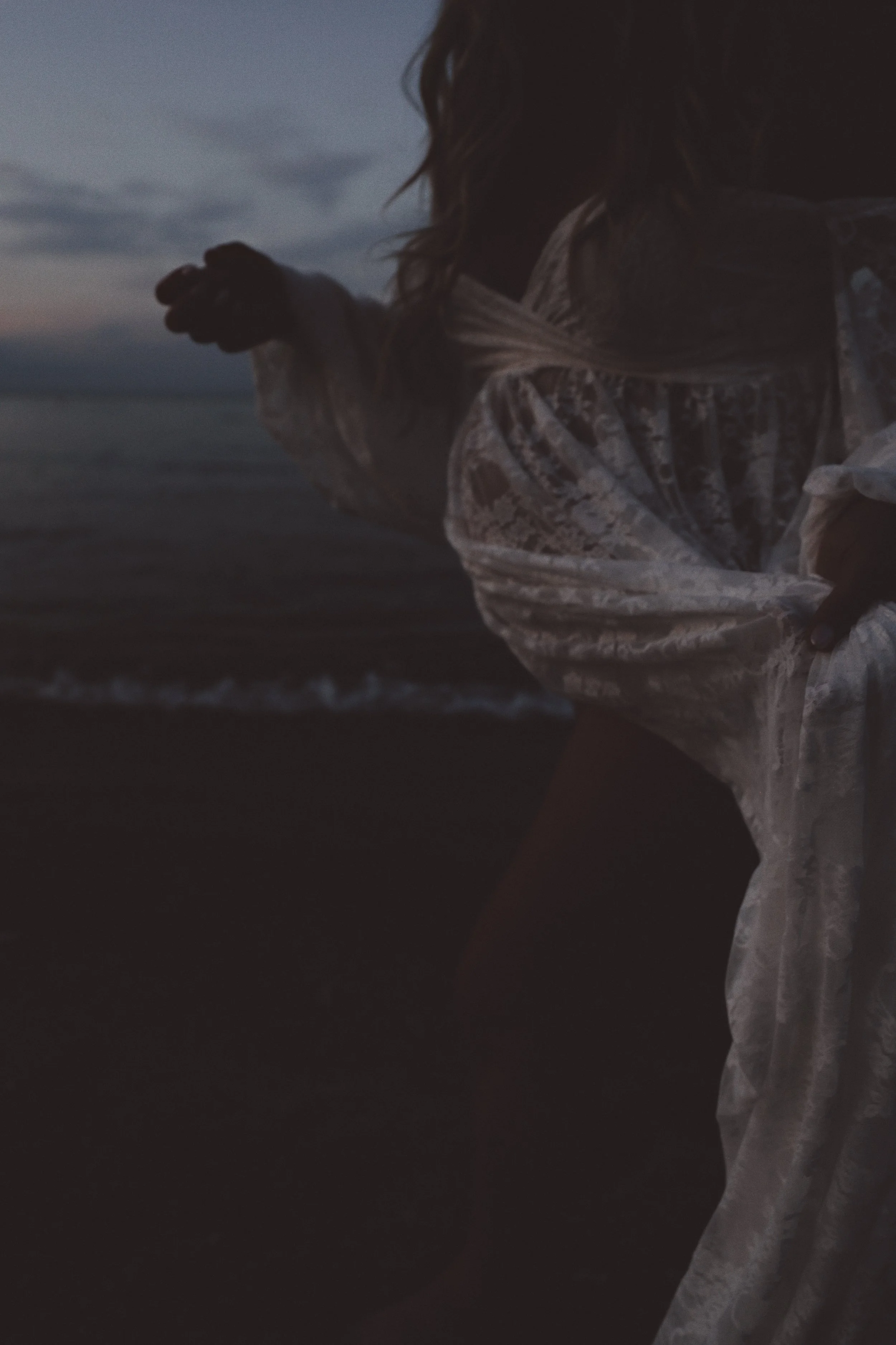 A woman at the beach during dusk, wearing a sheer, patterned dress with wavy hair, with the ocean and cloudy sky in the background.