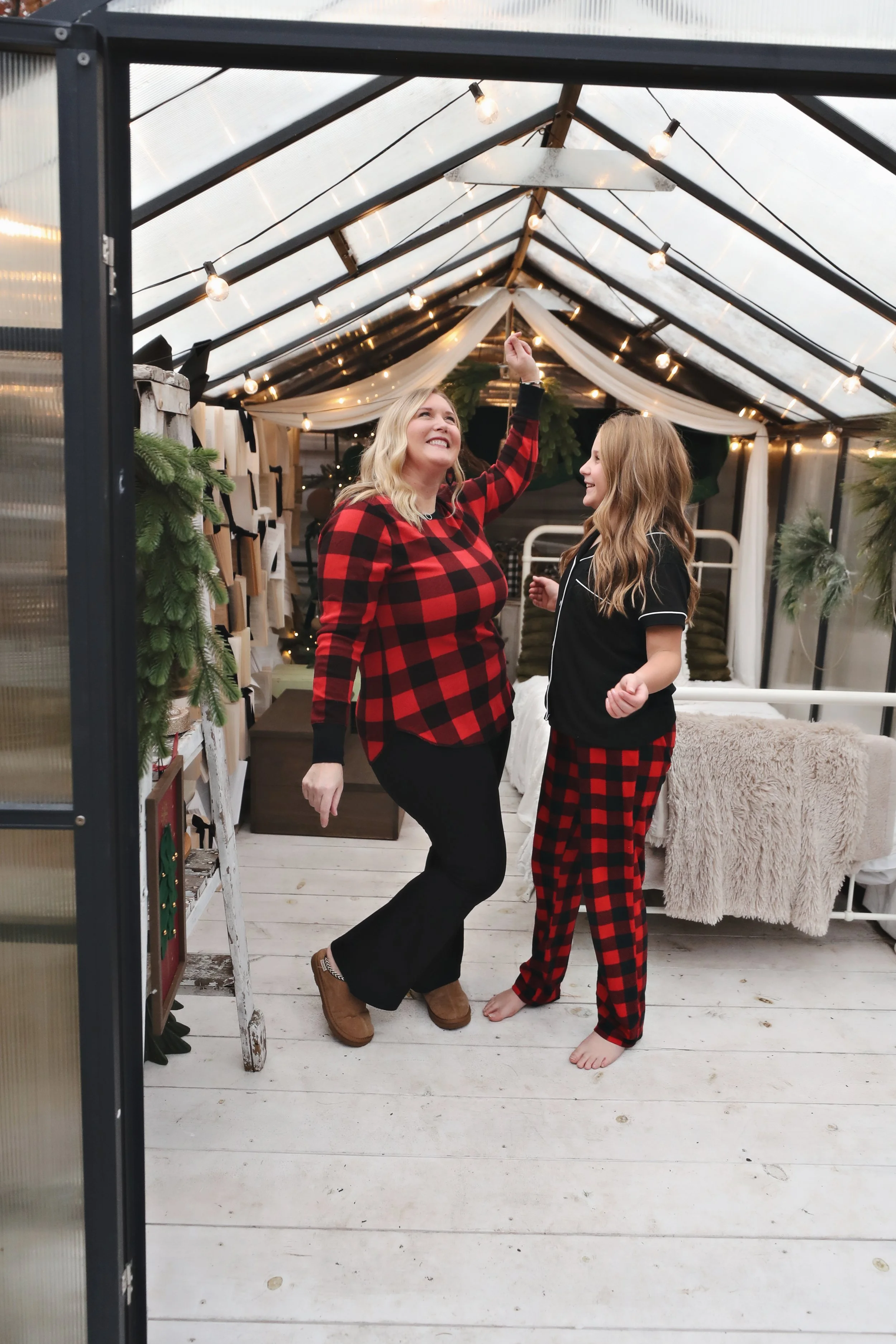 A woman and a girl dancing and smiling inside a glass greenhouse decorated with string lights and Christmas greenery, both wearing matching red and black plaid pajamas.