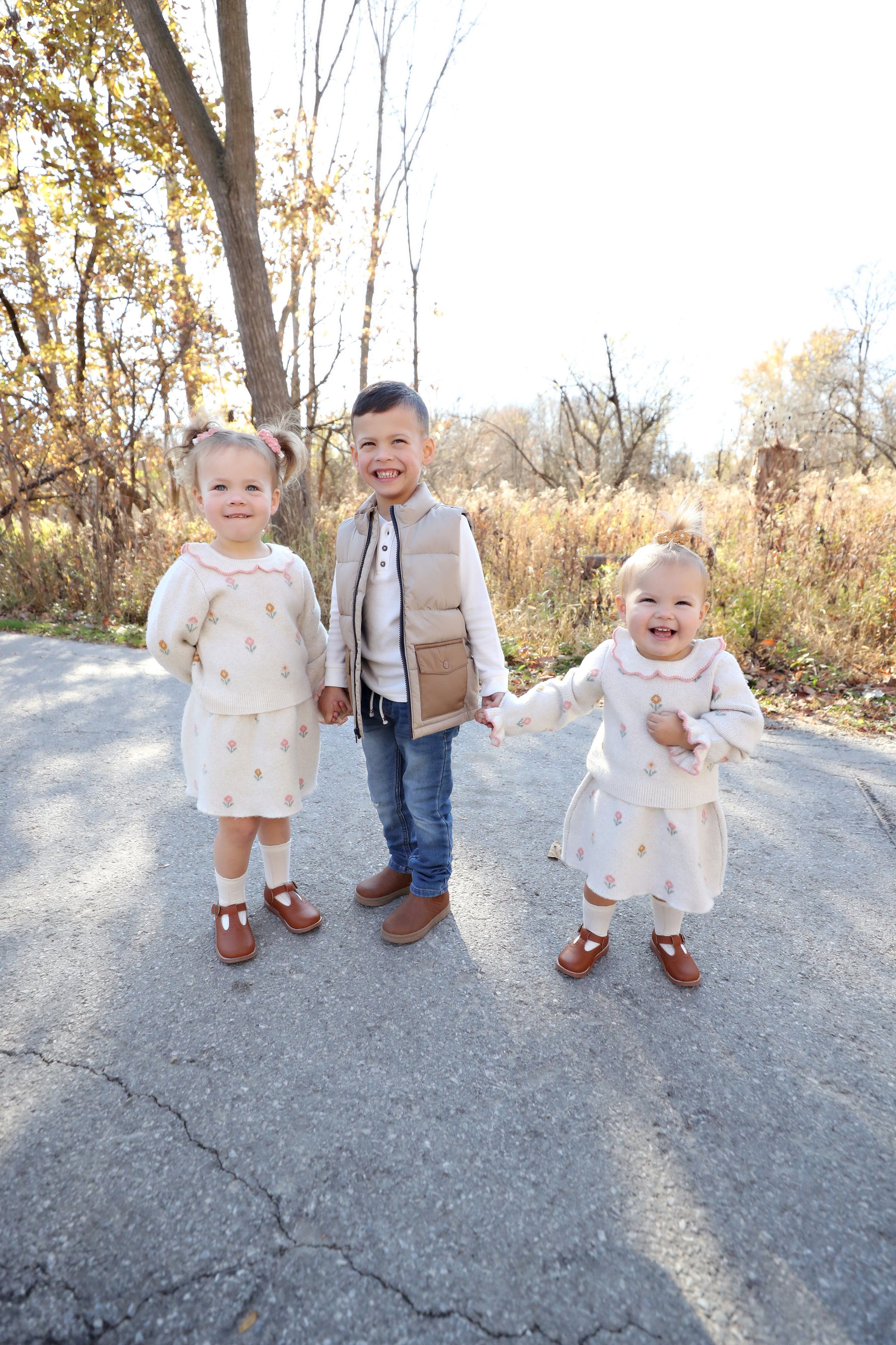 Three young children holding hands and smiling outdoors on a sunny day, with trees and autumn foliage in the background.