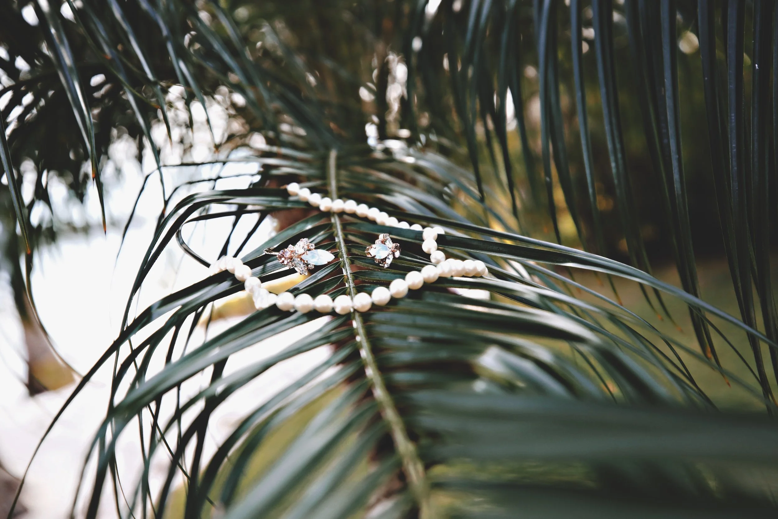 A pearl necklace and a pair of earrings with gemstones, resting on green palm leaves outdoors.