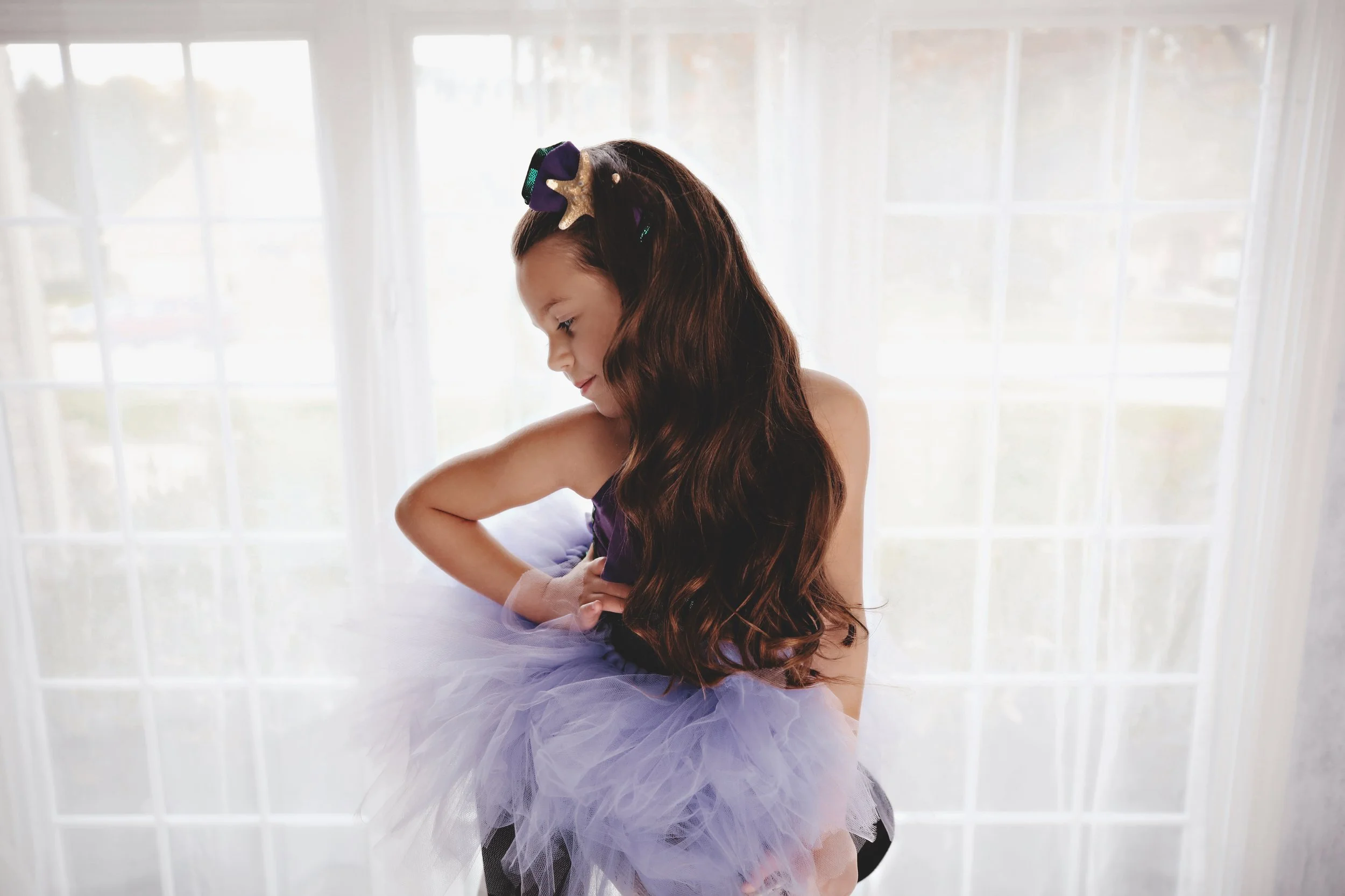 Young girl in a purple tutu dress and matching headband with star decorations, sitting in front of a large window with sheer curtains, looking down and smiling.