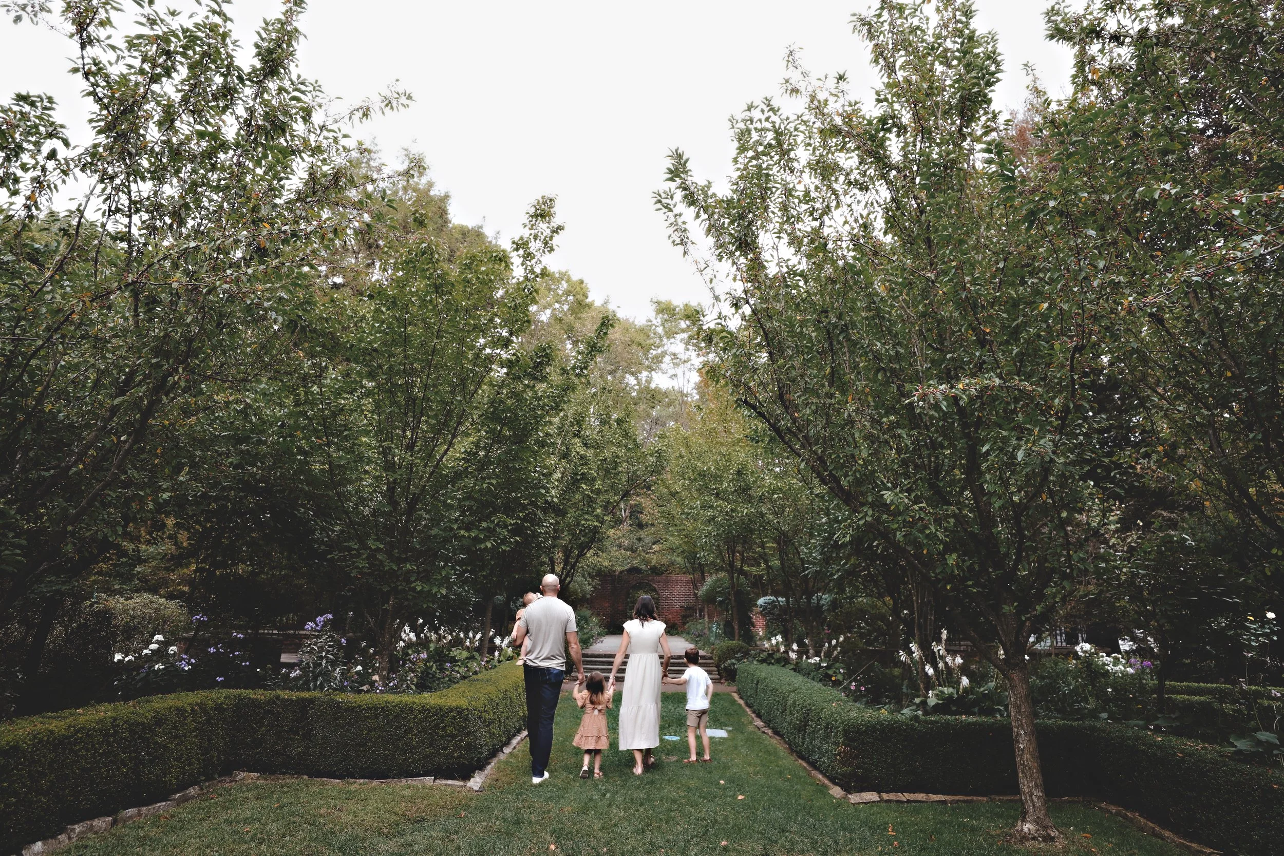 A family walking in a lush green garden with trimmed bushes and tall trees, during daytime.