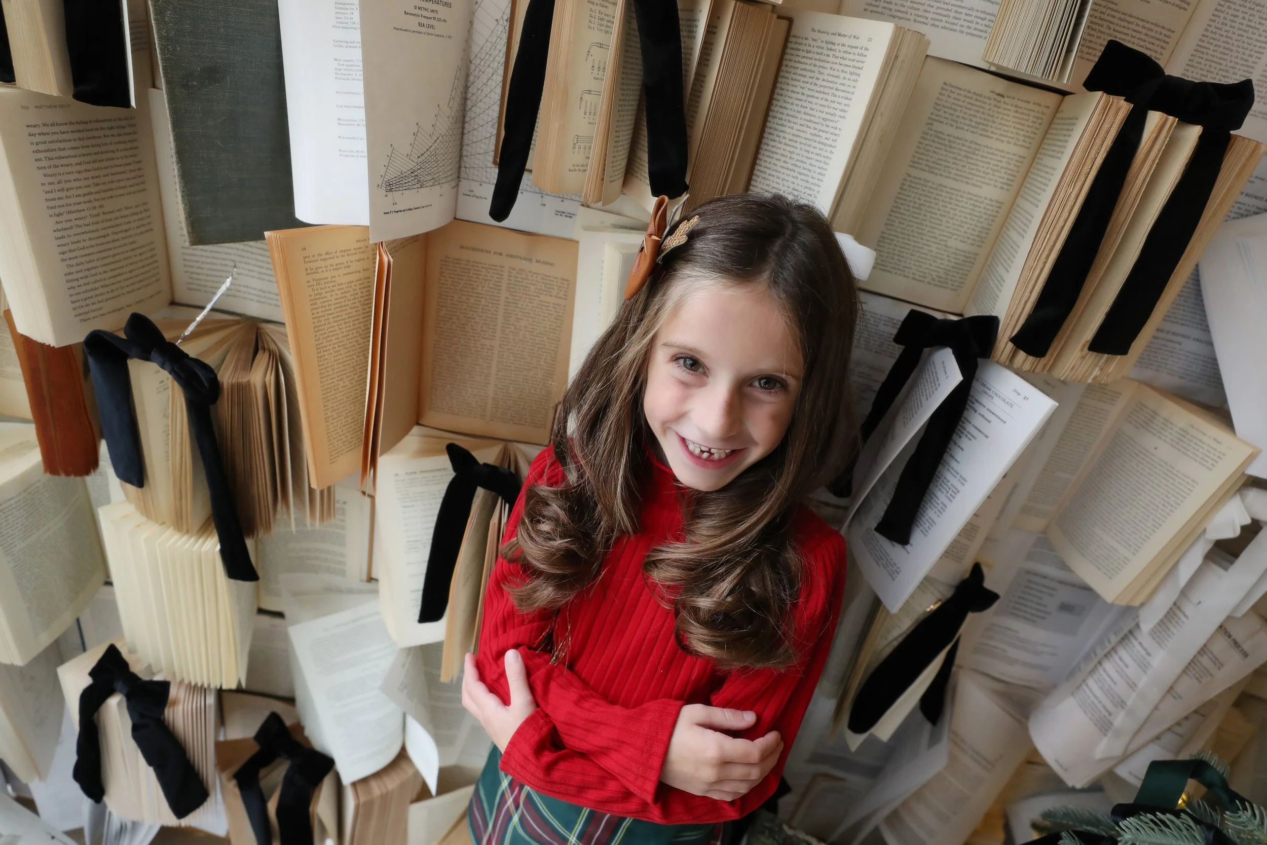 A young girl with long brown hair, wearing a red sweater and a plaid skirt, standing in front of a wall filled with open books with black ribbon markers, smiling at the camera.