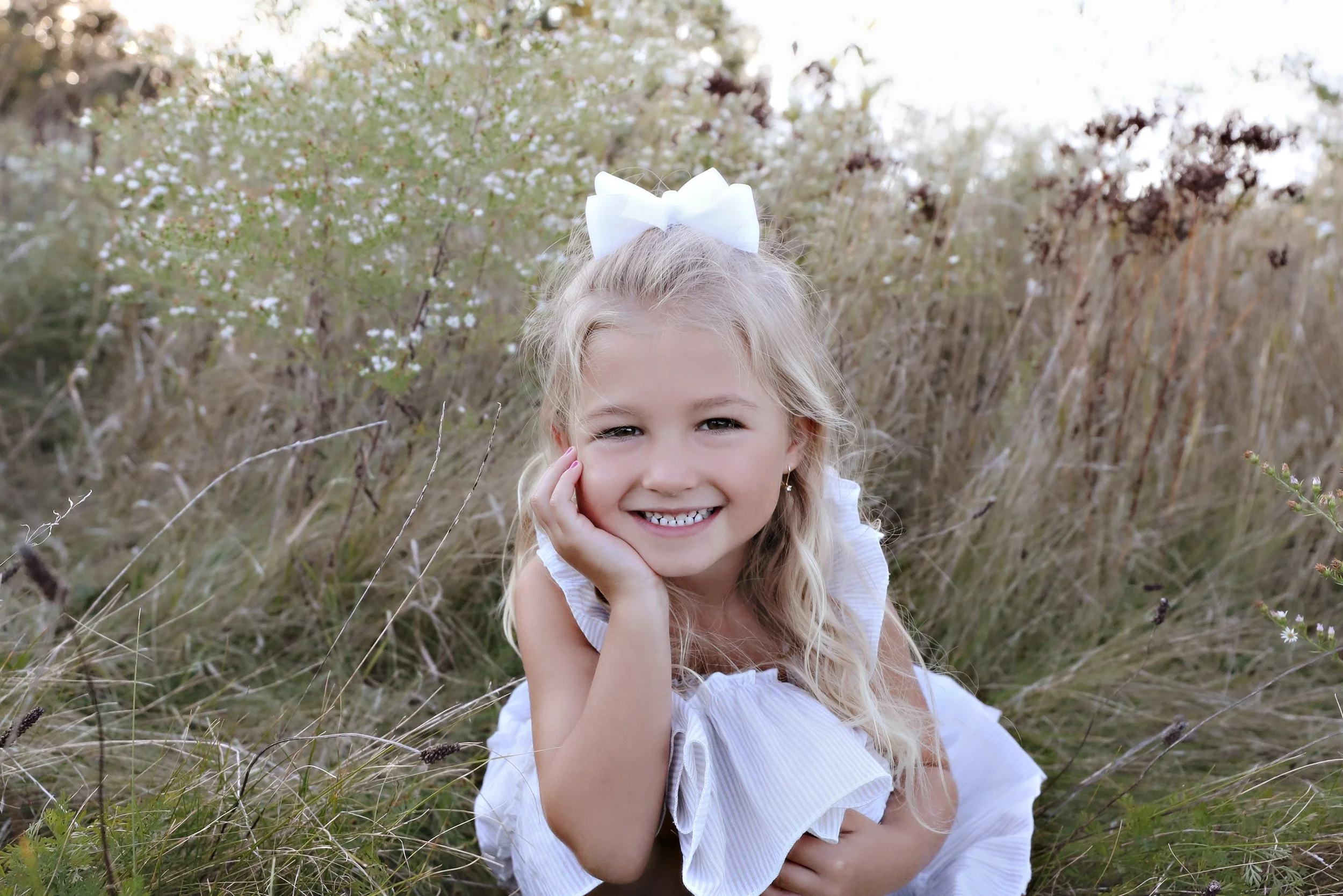 A young girl with long blonde hair, smiling and lying on her side in a grassy field, wearing a white dress with ruffled sleeves and a large white bow in her hair.