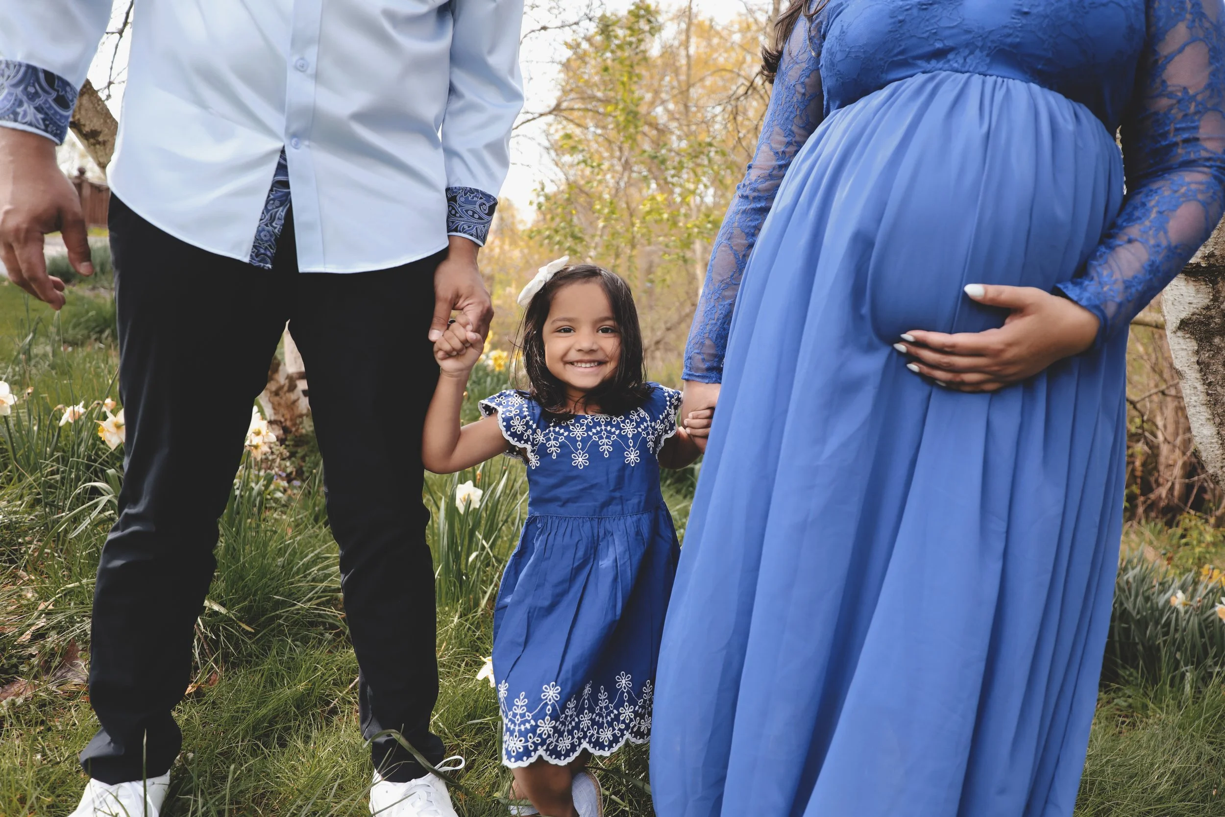 A happy young girl dressed in a blue dress holding hands with her pregnant mother, who is wearing a blue dress, and her father, wearing a white shirt and black pants, outdoors in a grassy area with trees and flowers.