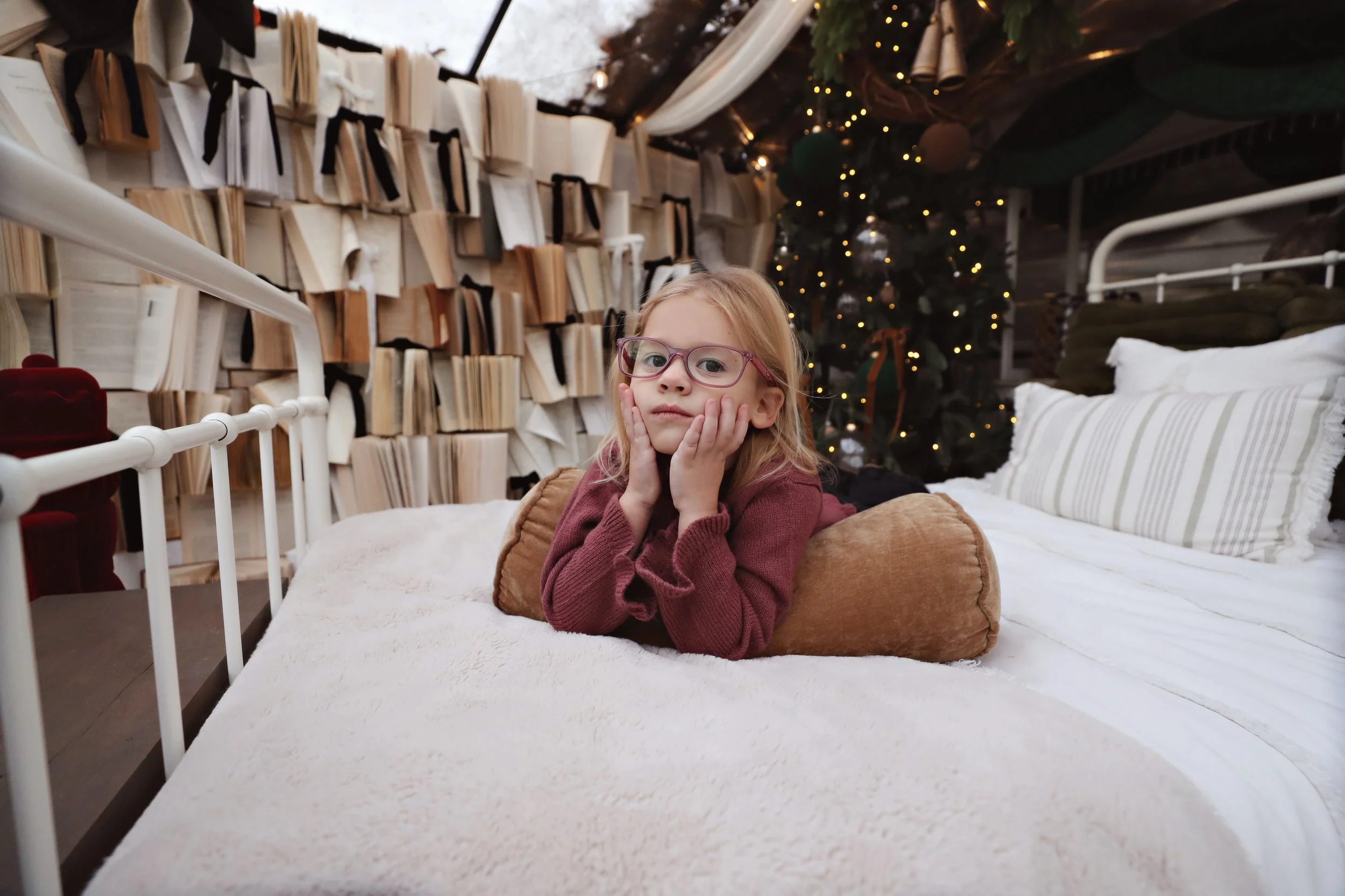 A young girl with red hair and glasses lying on her stomach on a bed, resting her head on her hands, in a cozy room decorated for Christmas with a Christmas tree and books on the wall behind her.