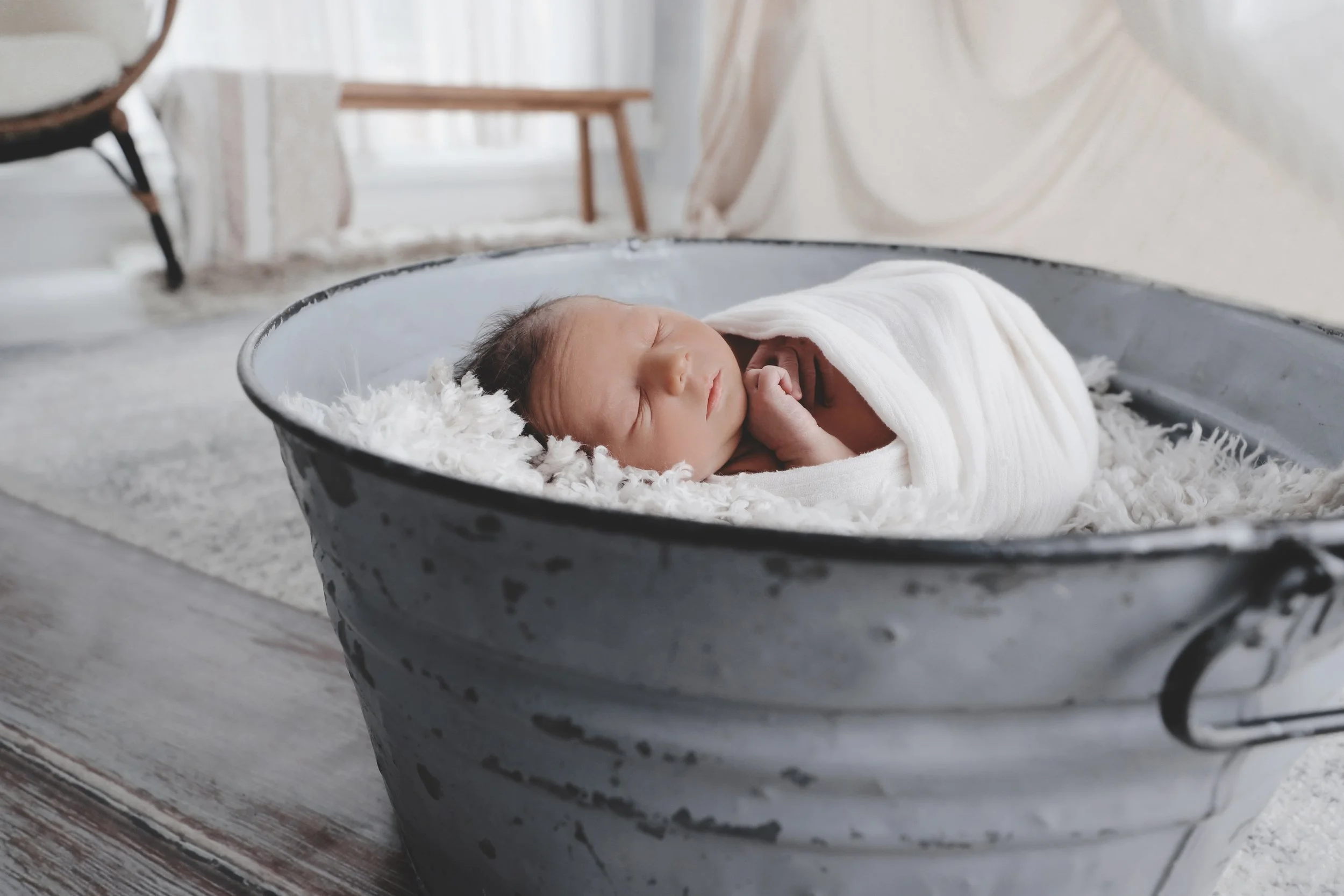 A newborn baby sleeping wrapped in a white blanket in a gray metal tub with fluffy white lining.