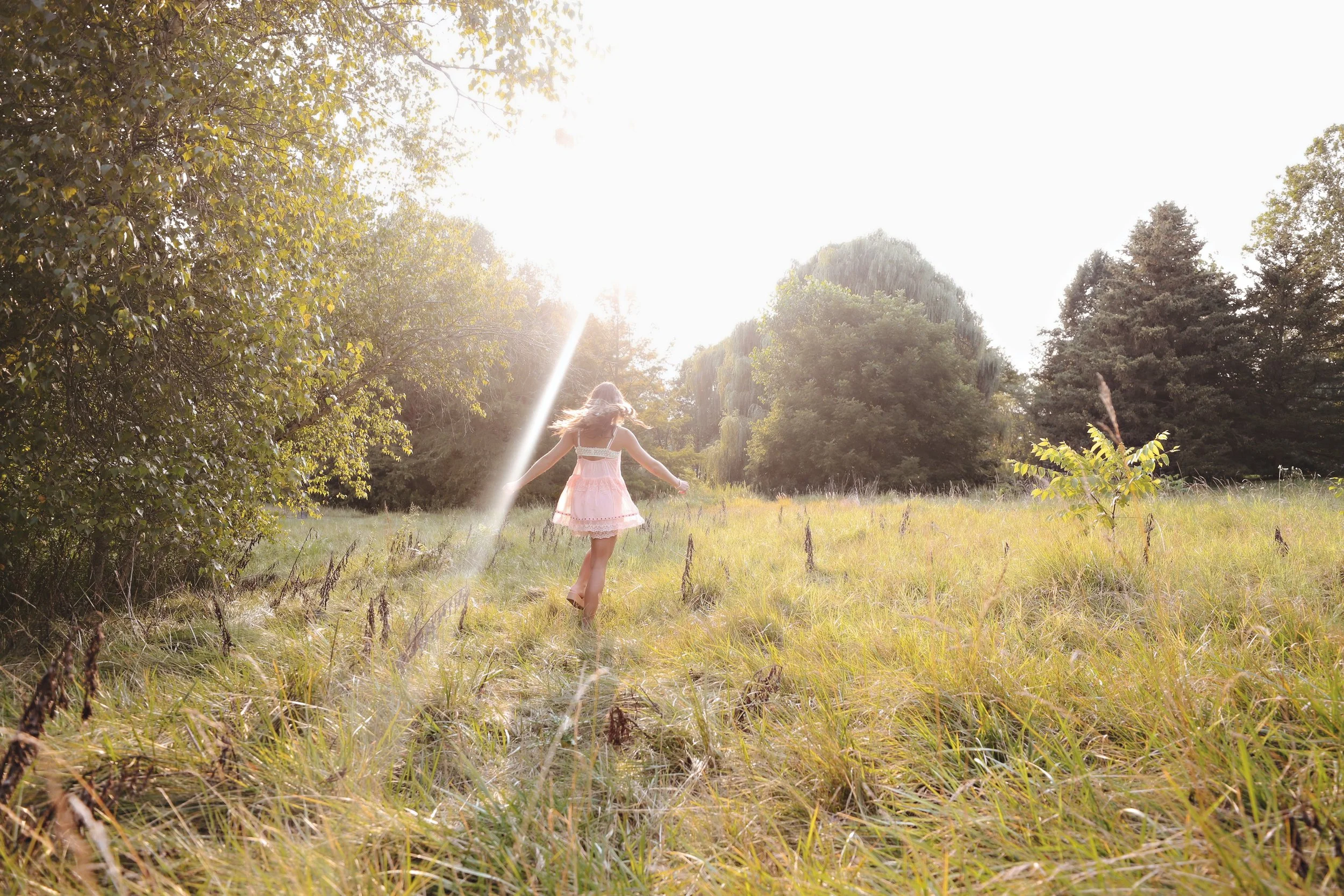 A girl in a pink dress running through a grassy field with trees in the background, sunlight shining brightly overhead.
