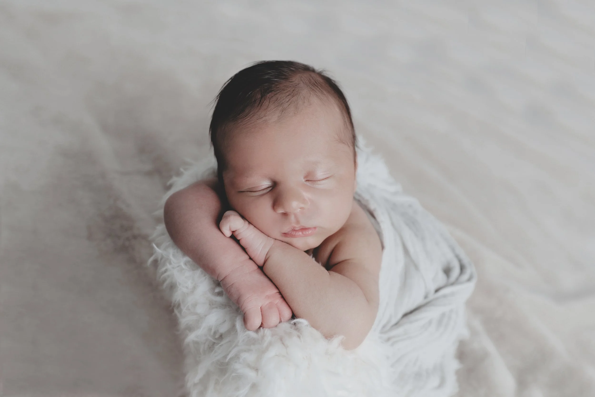Close-up of a sleeping newborn baby with dark hair, resting their head on one hand, lying on a soft, fluffy white blanket.