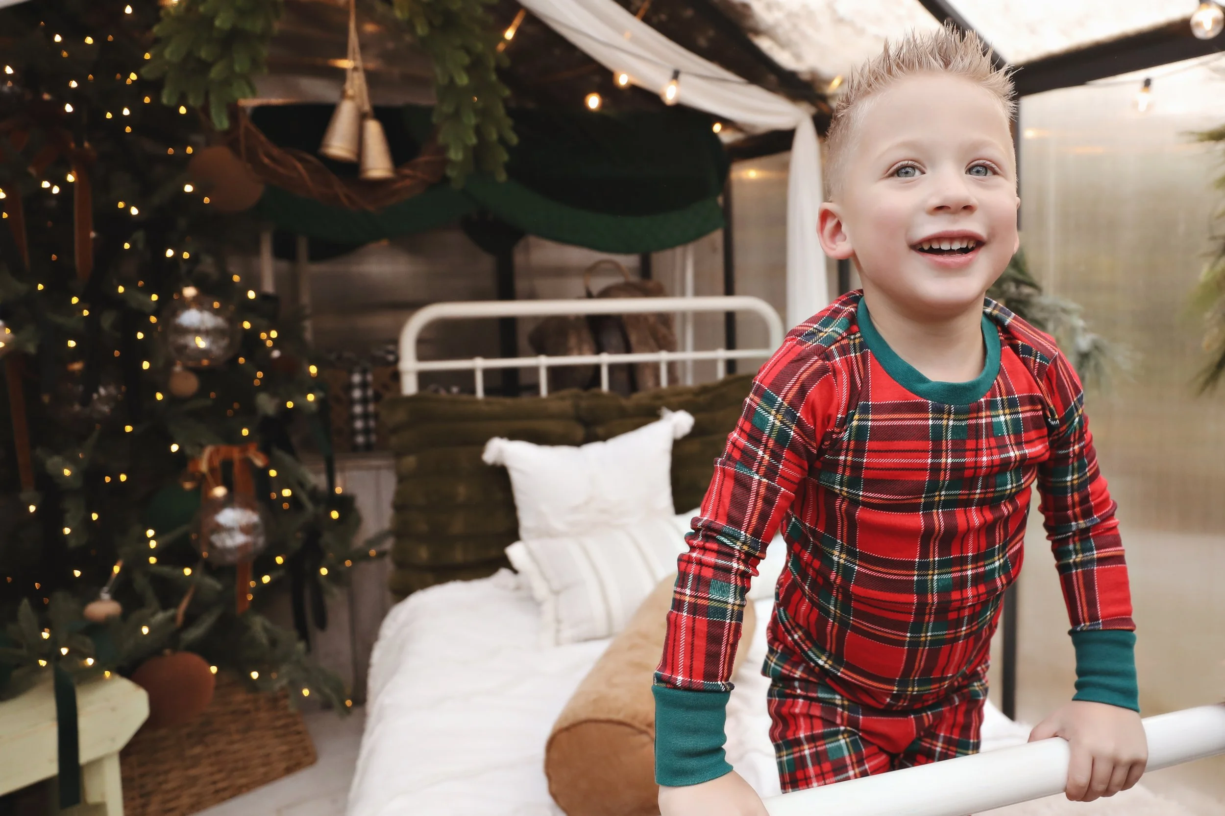Smiling young boy in plaid pajamas playing on a white bed in a decorated Christmas room with a Christmas tree and cushions.