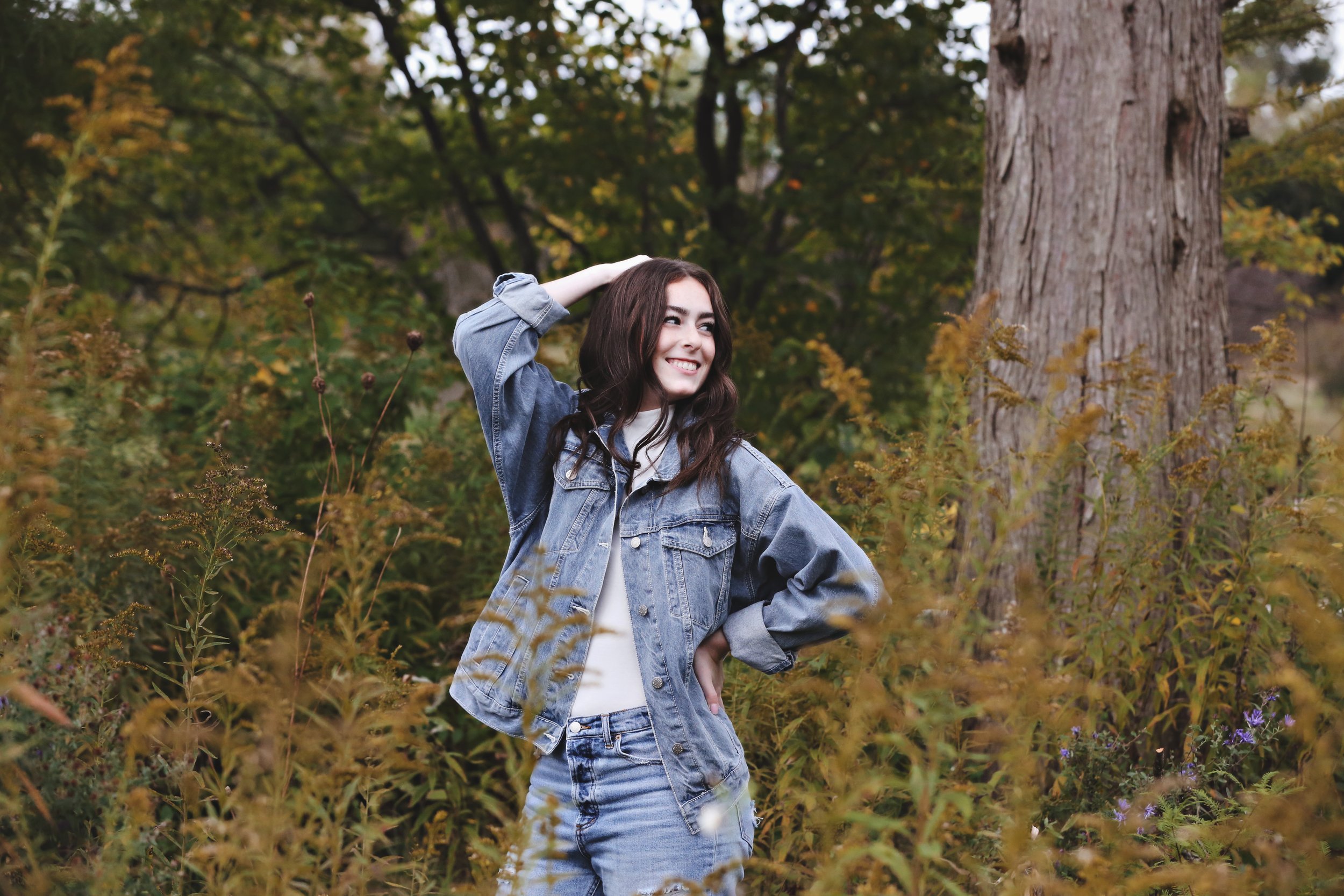 Young woman in denim jacket and jeans standing in a wooded area with trees and foliage, smiling and looking to the side.