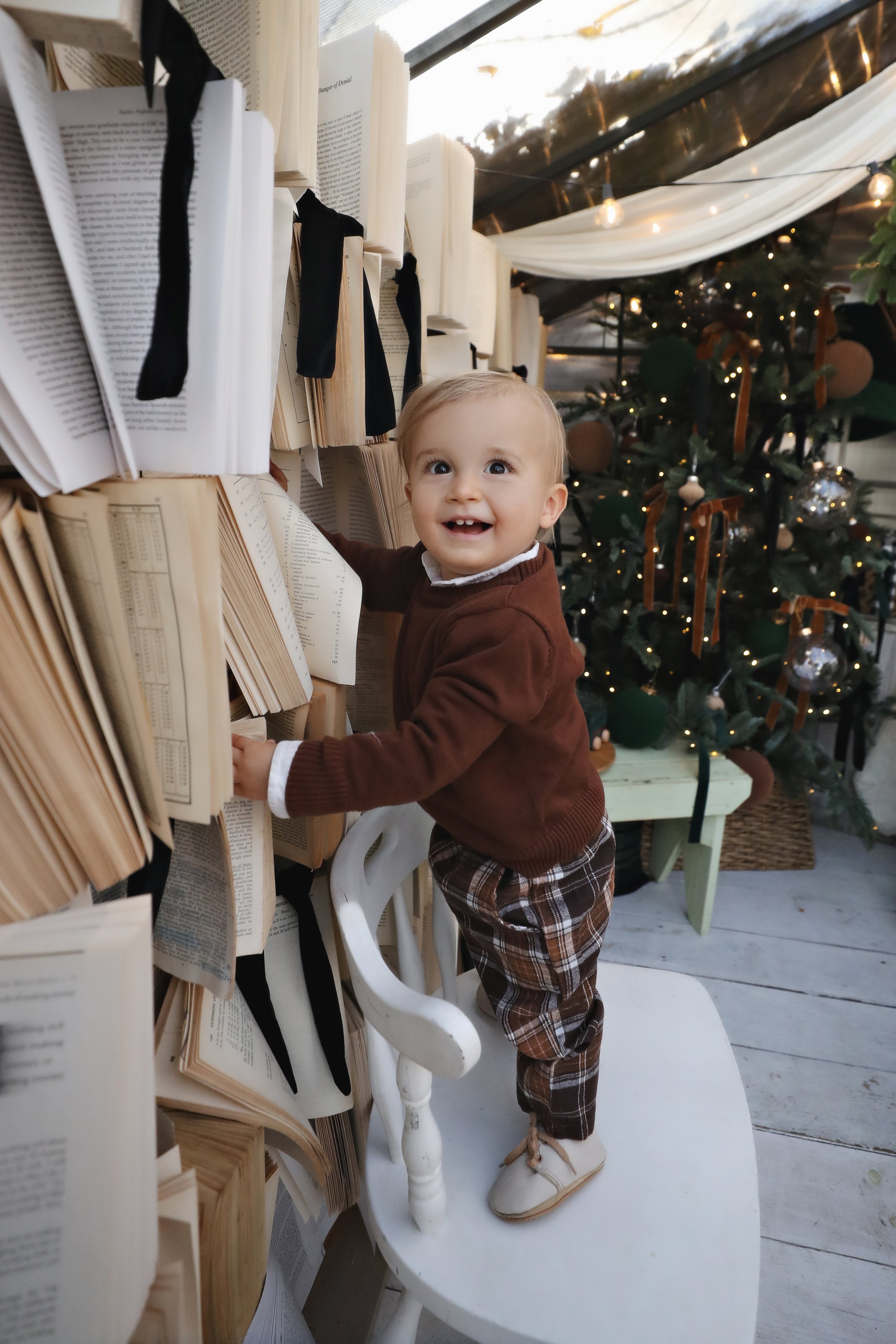 A young child standing on a white chair, reaching for an open book on a bookshelf with black ribbon bookmarks, next to a decorated Christmas tree with ornaments and lights.
