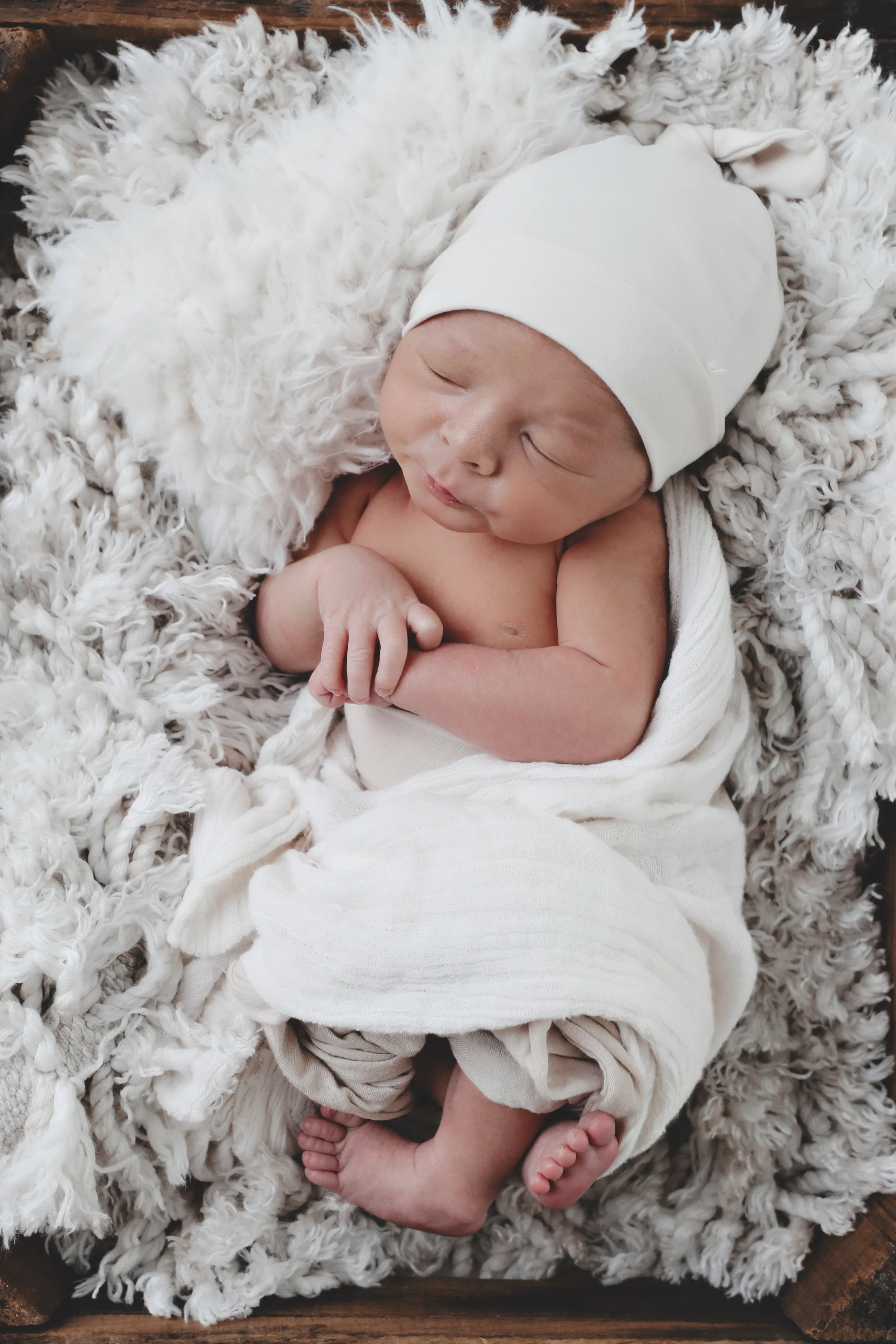A newborn baby sleeping wrapped in white cloth, wearing a white hat, lying on a fluffy white blanket.