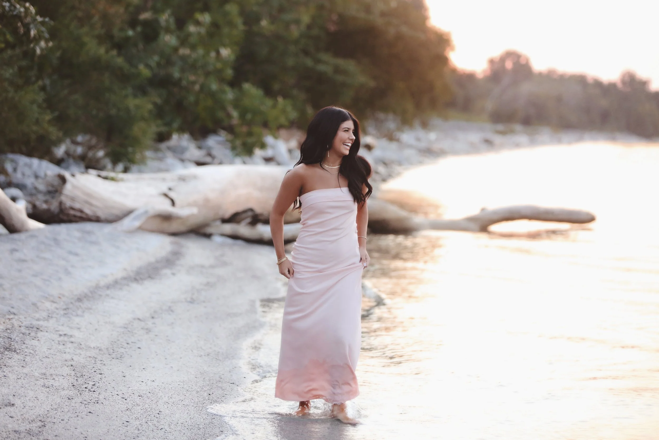 Woman in a strapless, light pink dress smiling and walking along a sandy beach at sunset, with trees and driftwood in the background.