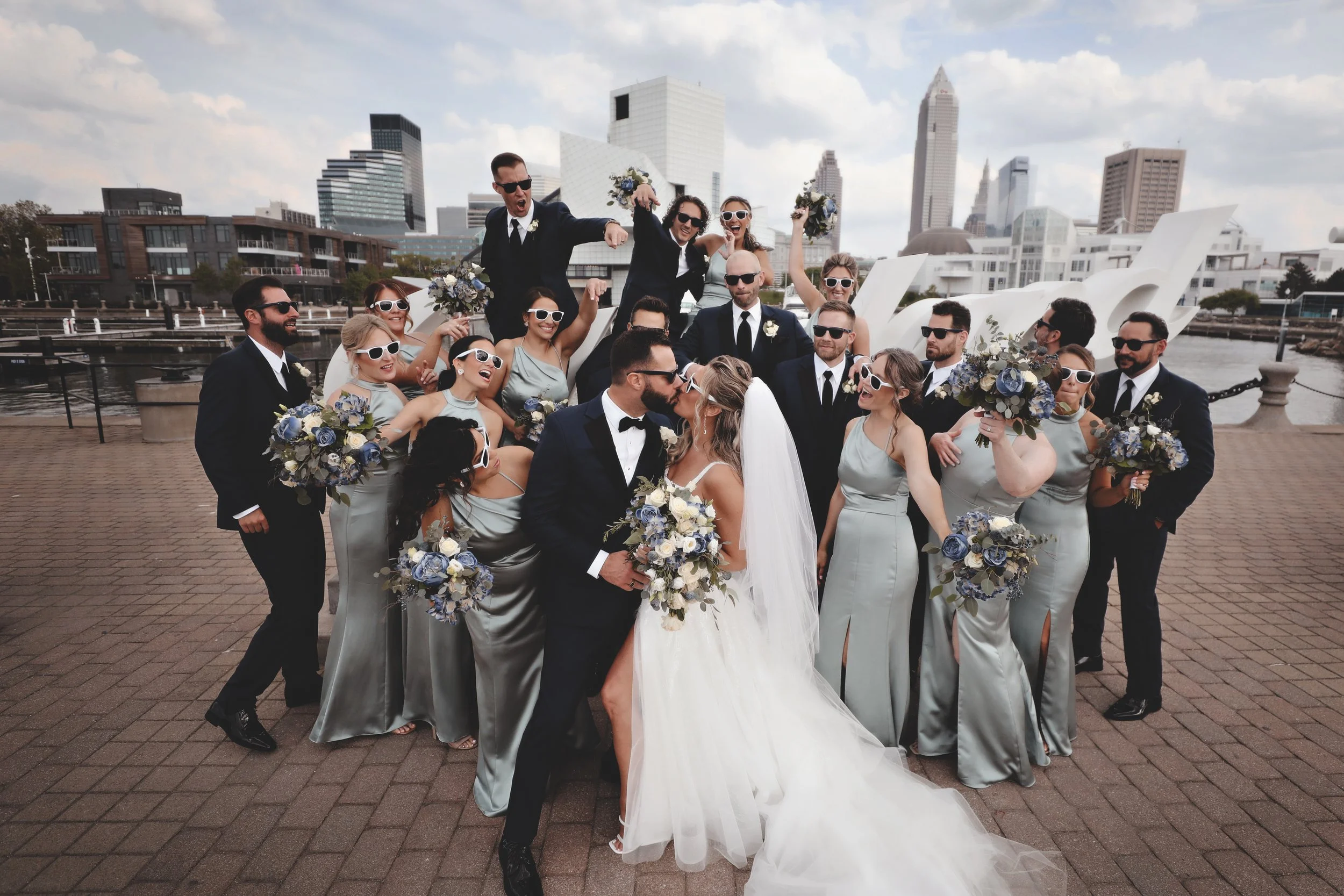 A wedding party of 21 people, including bride and groom, posing outdoors with city skyline in the background. The group is dressed in formal attire, with men in suits and women in matching light gray dresses holding bouquets. Some members are wearing