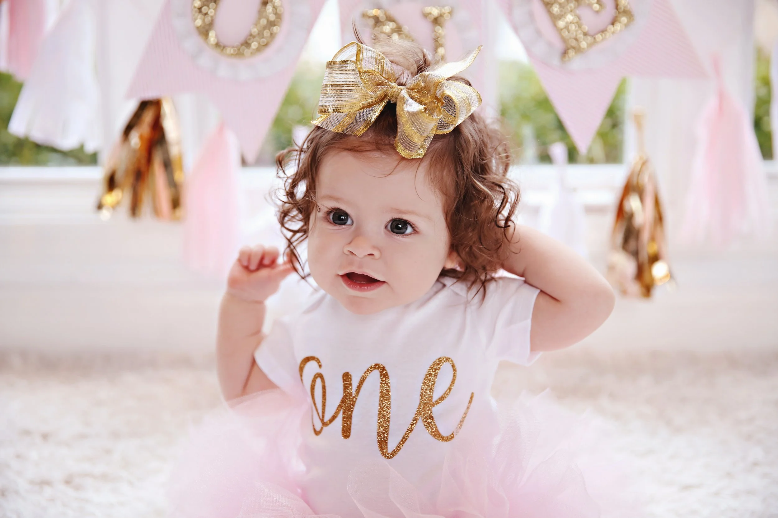 A young girl with curly brown hair wearing a gold bow headband, a white shirt with the word "one" in glittery gold, and a pink tutu, at a first birthday celebration with pink and gold decorations.