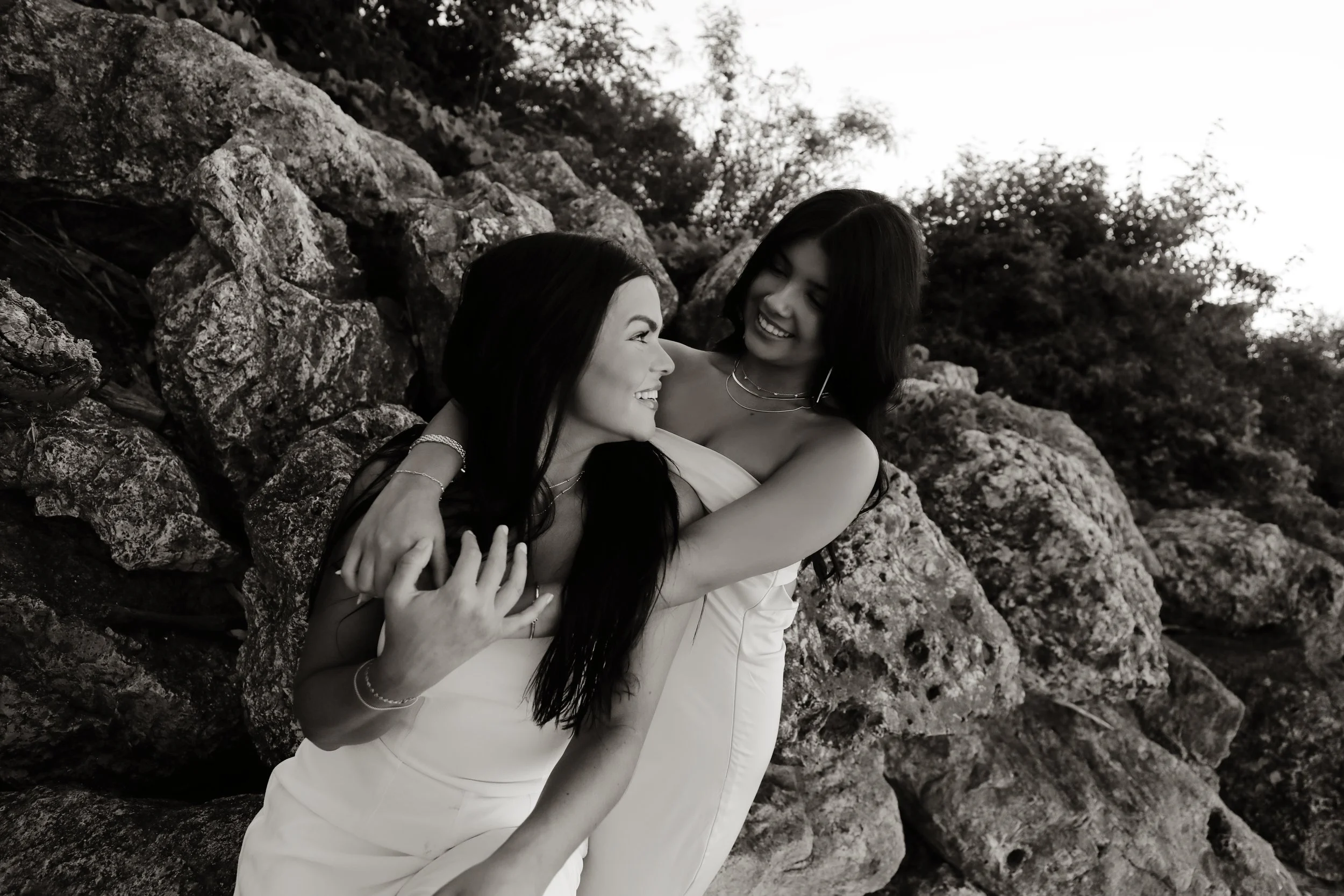 Two women smiling and hugging outdoors near rocks with trees in the background, in black and white.