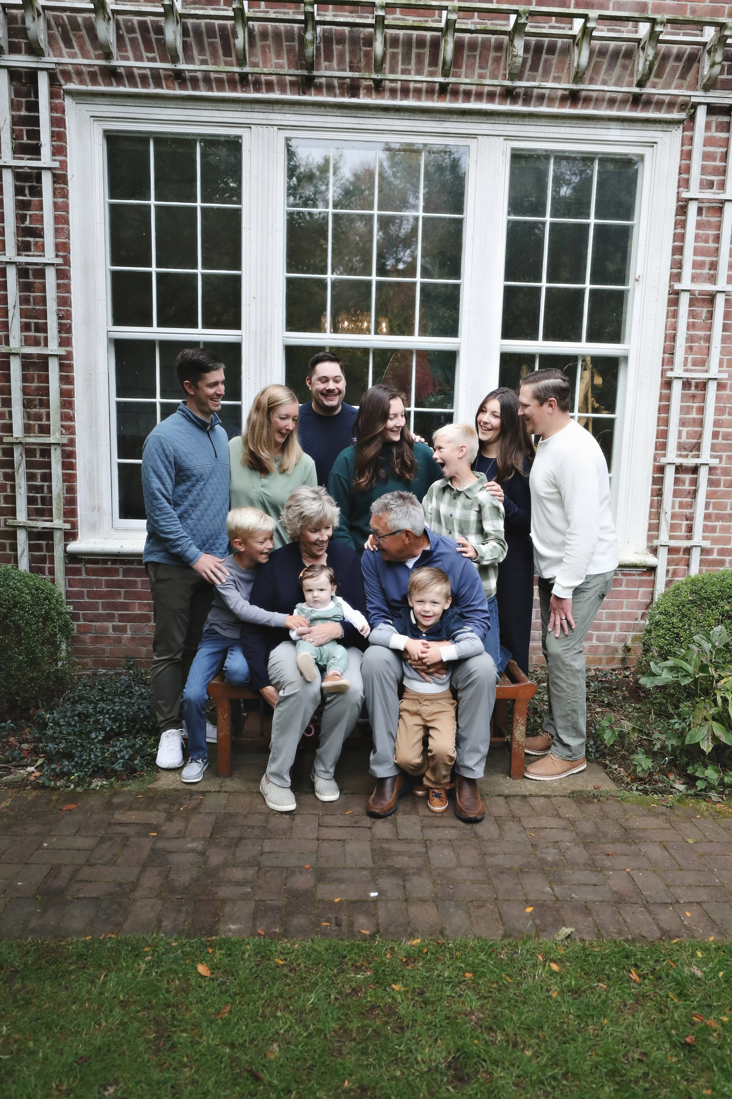 A large family gathering outdoors in front of a big window on a brick house, smiling and laughing together.