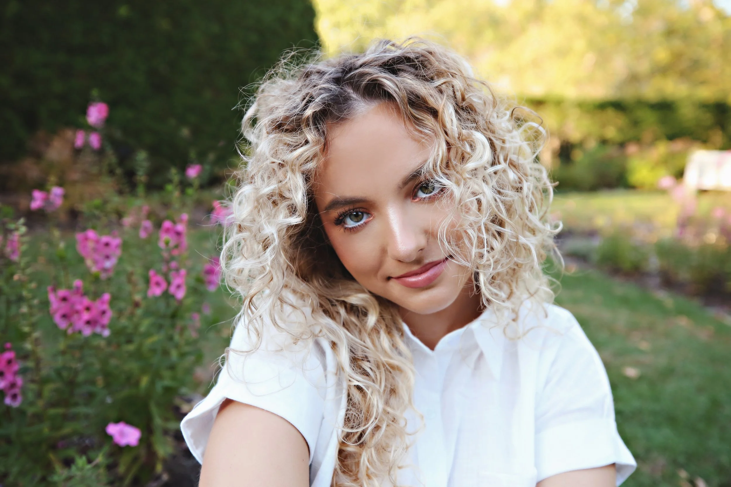 Young woman with curly blonde hair, blue eyes, wearing a white shirt, smiling in a garden with pink flowers and green foliage.
