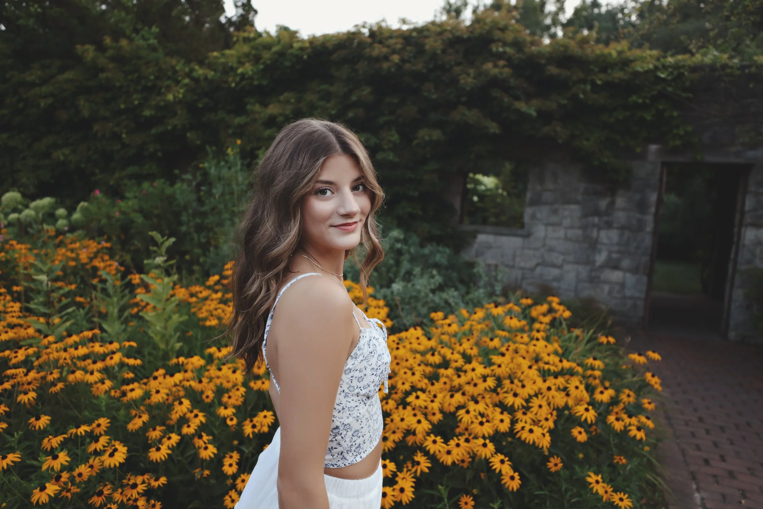 A young woman with long wavy brown hair standing in front of a garden with yellow flowers, wearing a white patterned sleeveless top and white pants, smiling softly at the camera.