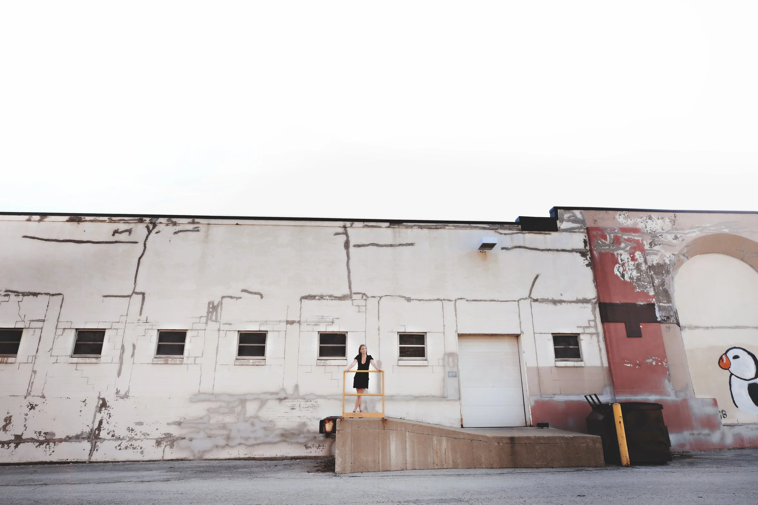 A woman standing behind a yellow railing on a concrete platform in front of a white, weathered building with small windows and graffiti art, including a cartoonish bird, on the wall.