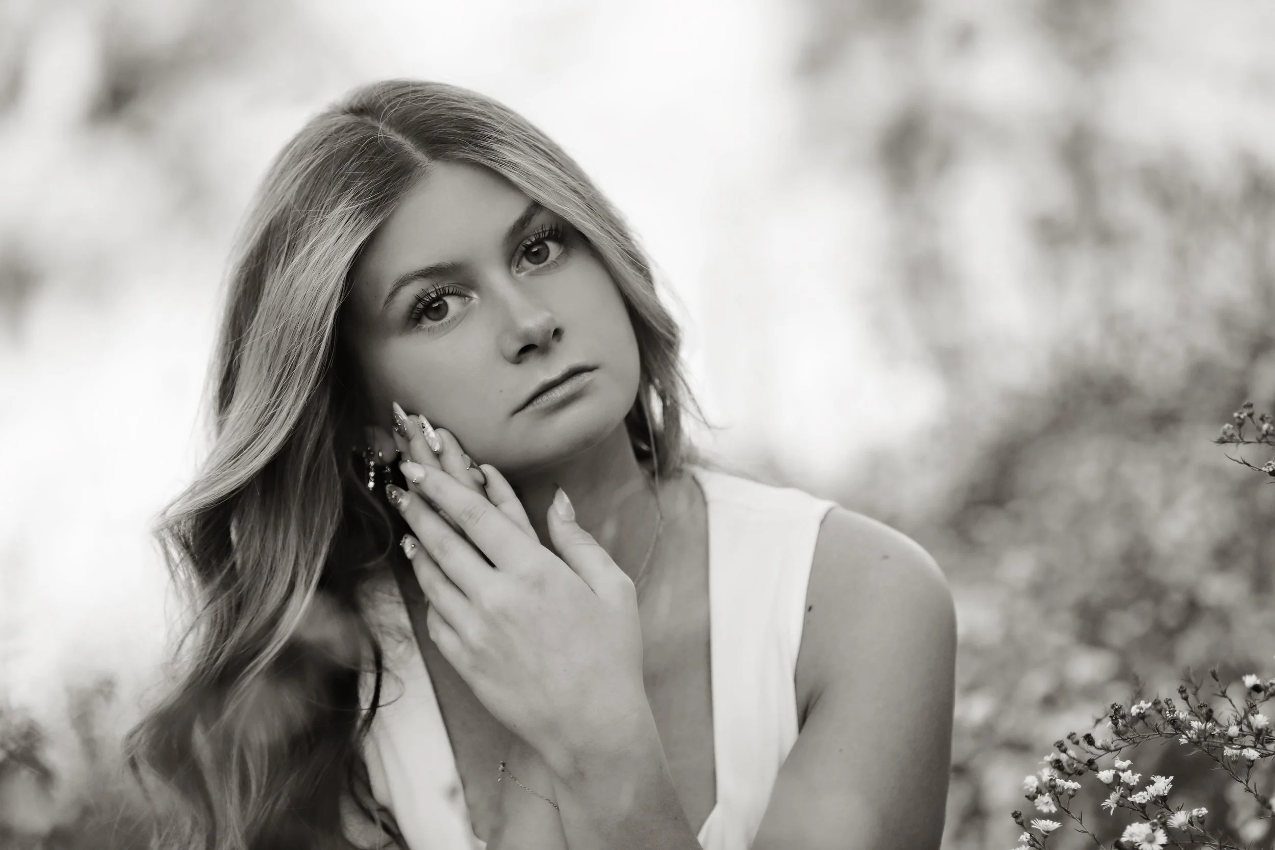 Black and white photo of a young woman with long wavy hair, looking thoughtfully at the camera, touching her face with her right hand, outdoors with blurred flowers in the background.
