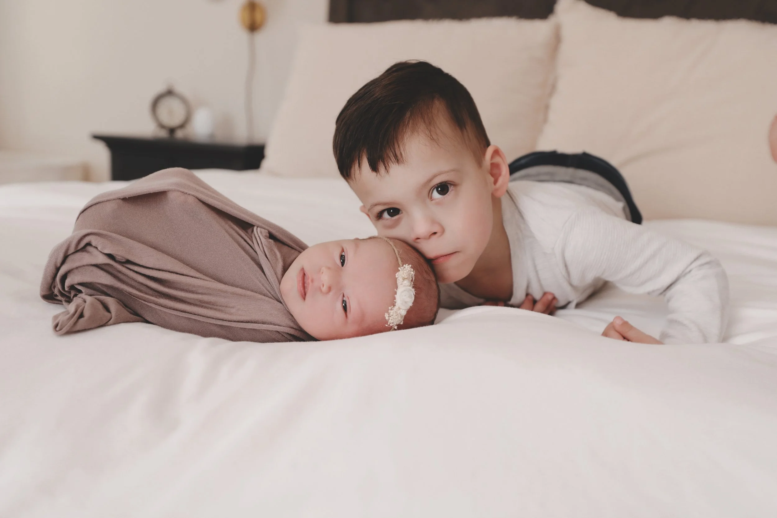 Young boy and newborn baby lying on a bed, with the boy gently resting his face on the baby's head, both facing the camera.