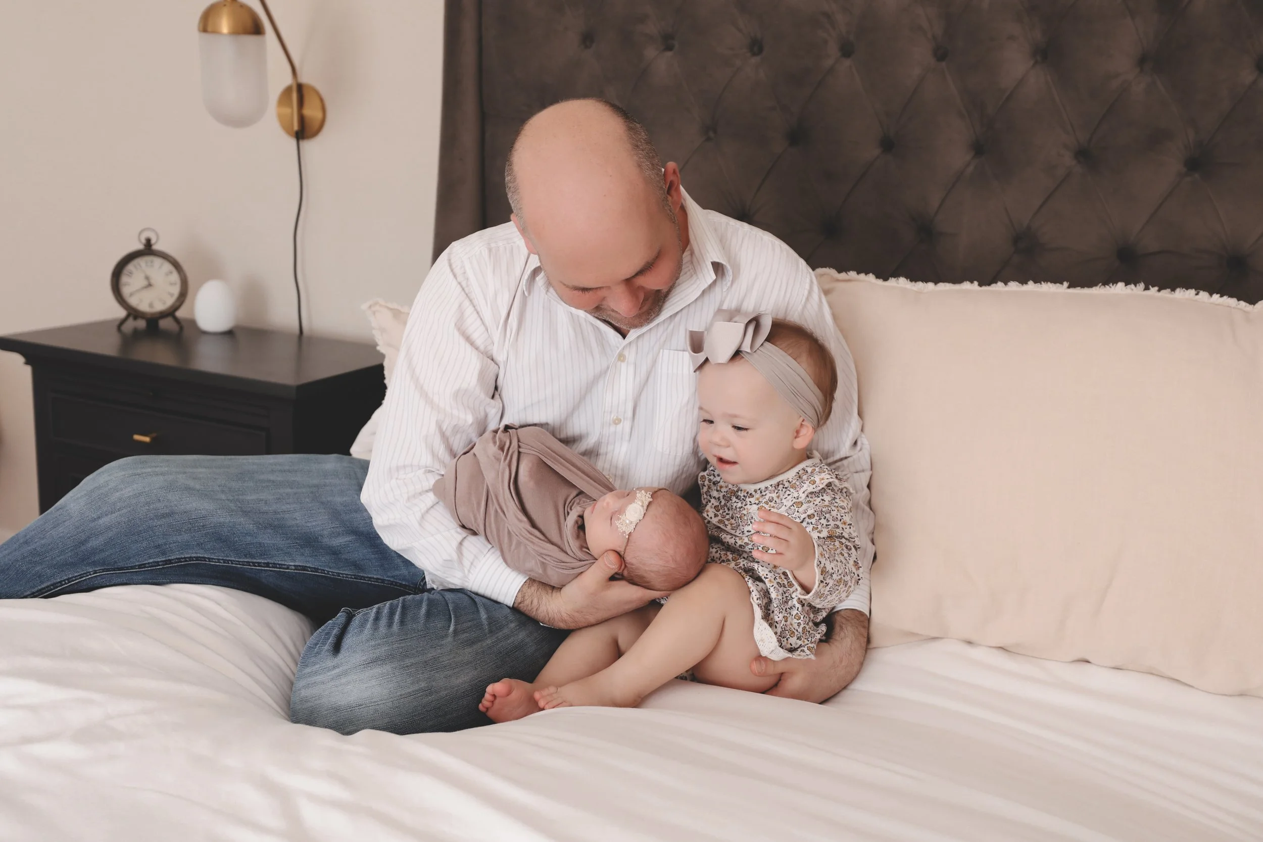 A man, likely a father, sitting on a bed holding a newborn baby wrapped in a blanket, while a young girl, probably a sibling, sits beside them, touching the baby and looking at him with curiosity and affection.