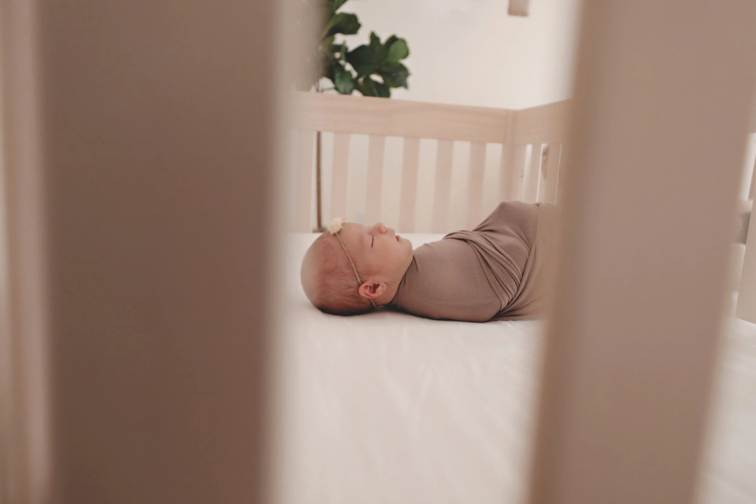 A baby sleeping on a bed, seen through white crib slats with a plant in the background.