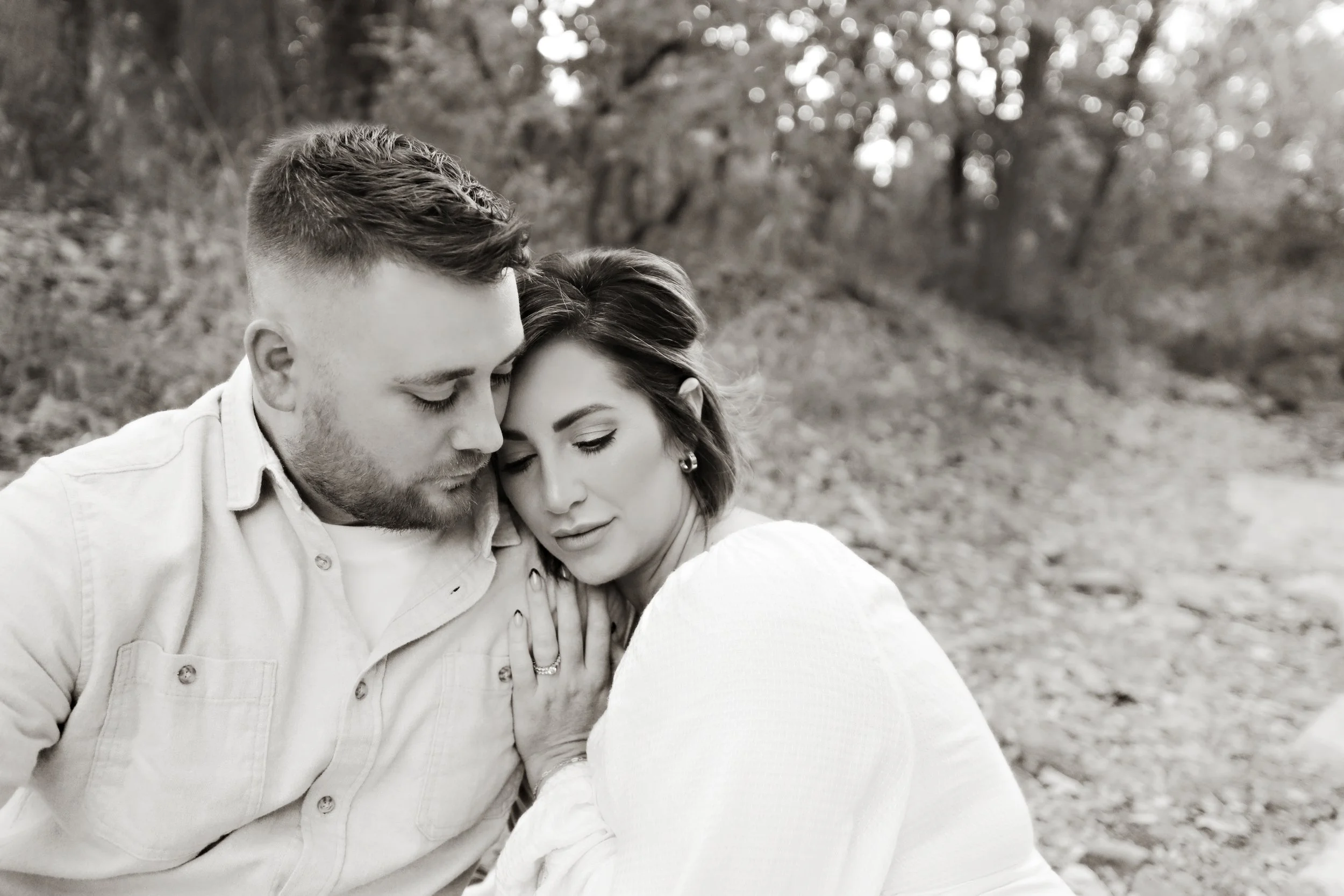 A black and white photo of a couple holding each other in an outdoor setting with trees in the background, their foreheads touching and eyes closed.