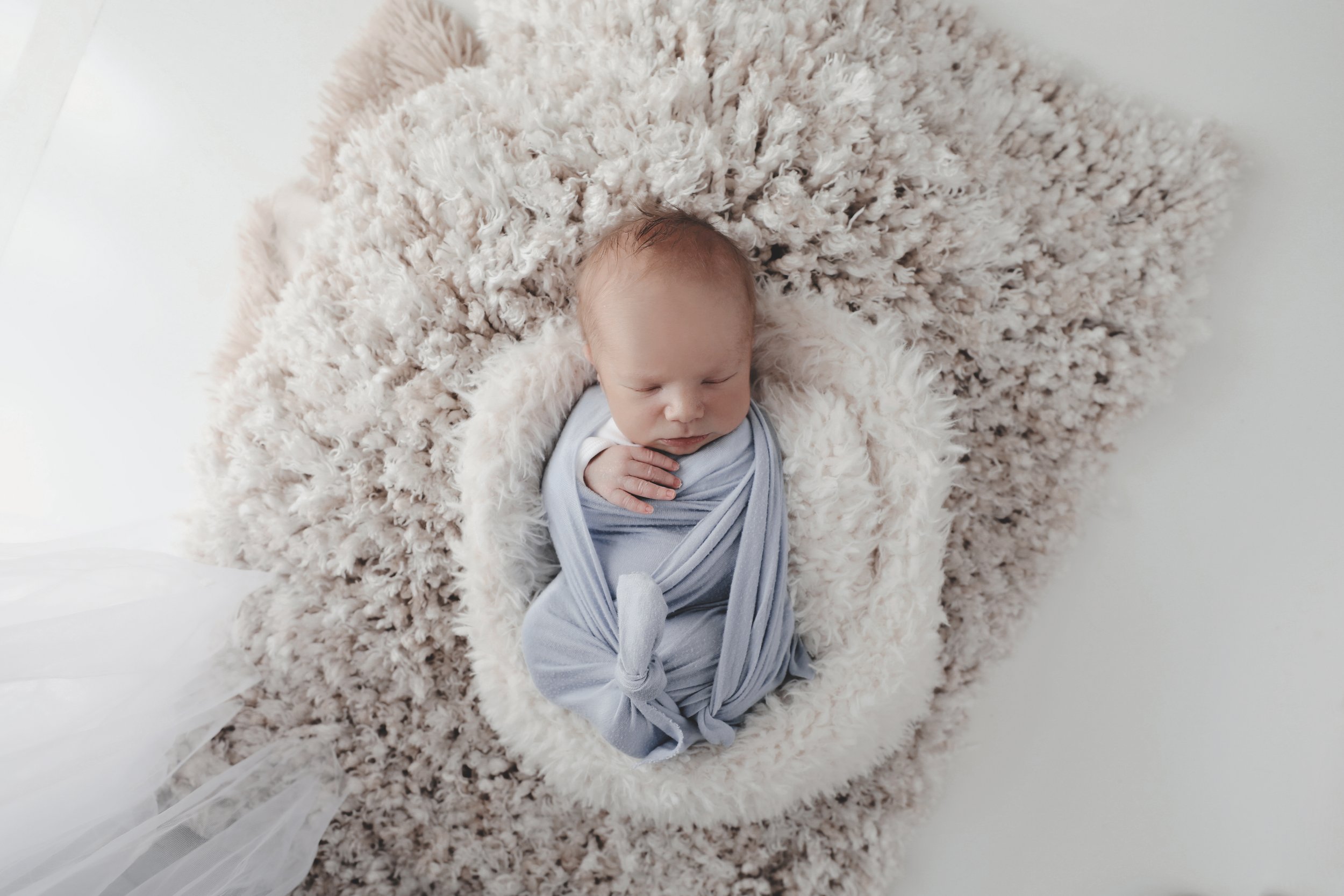 A sleeping newborn baby wrapped in a light gray swaddle, lying on a fluffy, cream-colored rug.