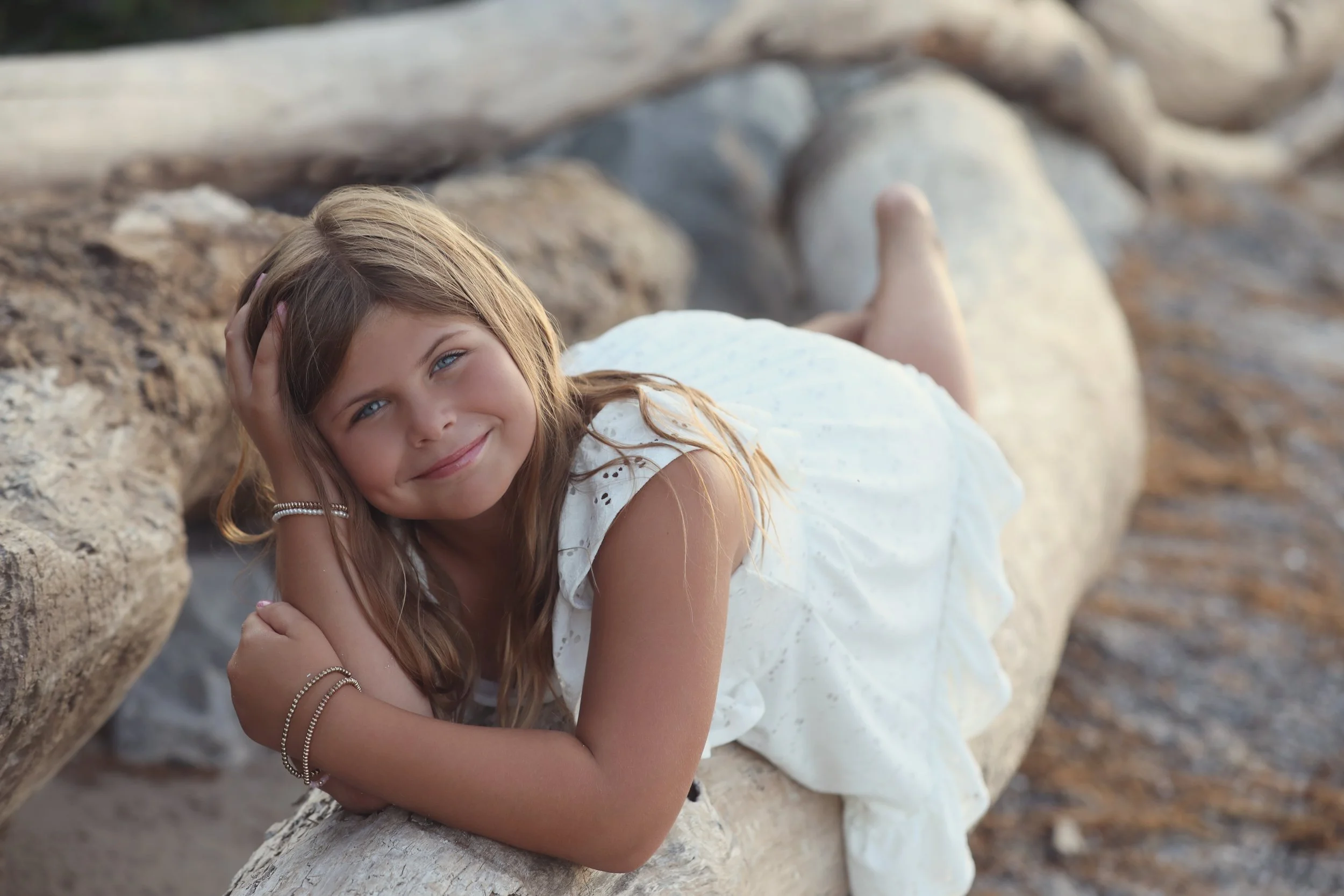 Young girl with long light brown hair lying on a large driftwood log at a beach, smiling at the camera with her head resting on her hand, wearing a white dress and jewelry.